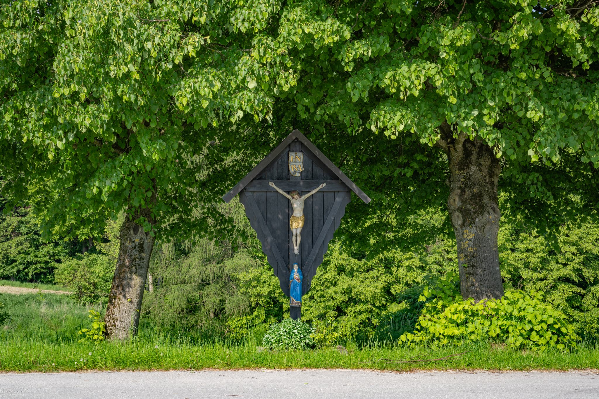 Traditionelles Flurkreuz mit Kruzifix und Marienstatue in Friesing, Reischach, Altötting, Oberbayern. Zwischen zwei Bäumen am Wegesrand.