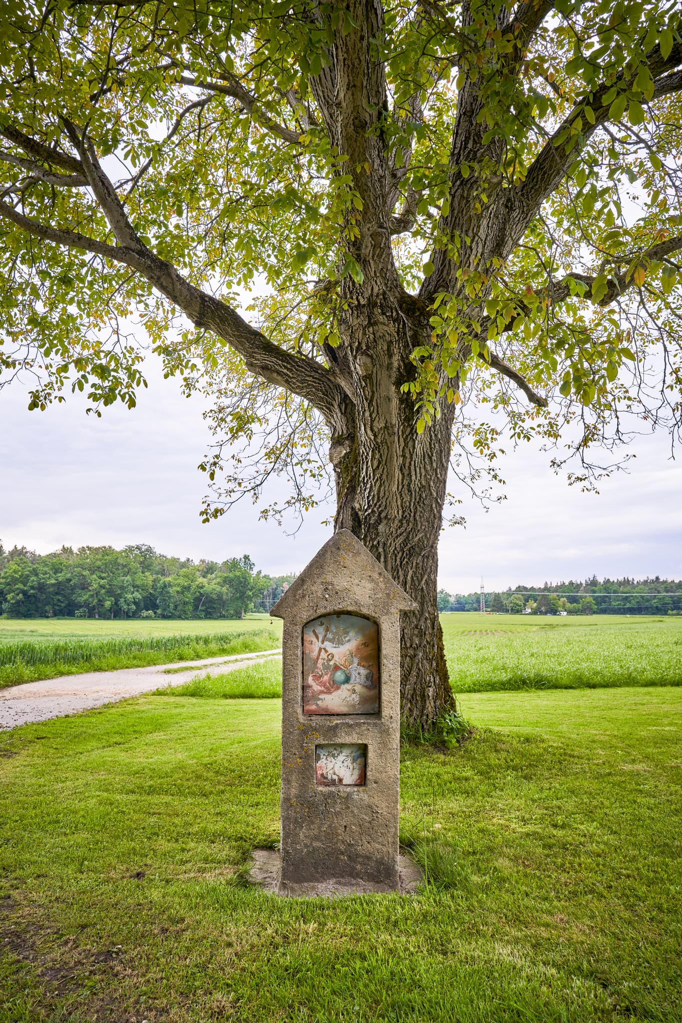 Historischer Bildstock am Feldweg bei Höresham, Burgkirchen, Altötting, Oberbayern. Grüne Wiesen, großer Baum im Hintergrund. Ländliche Schönheit Inn-Salzach.