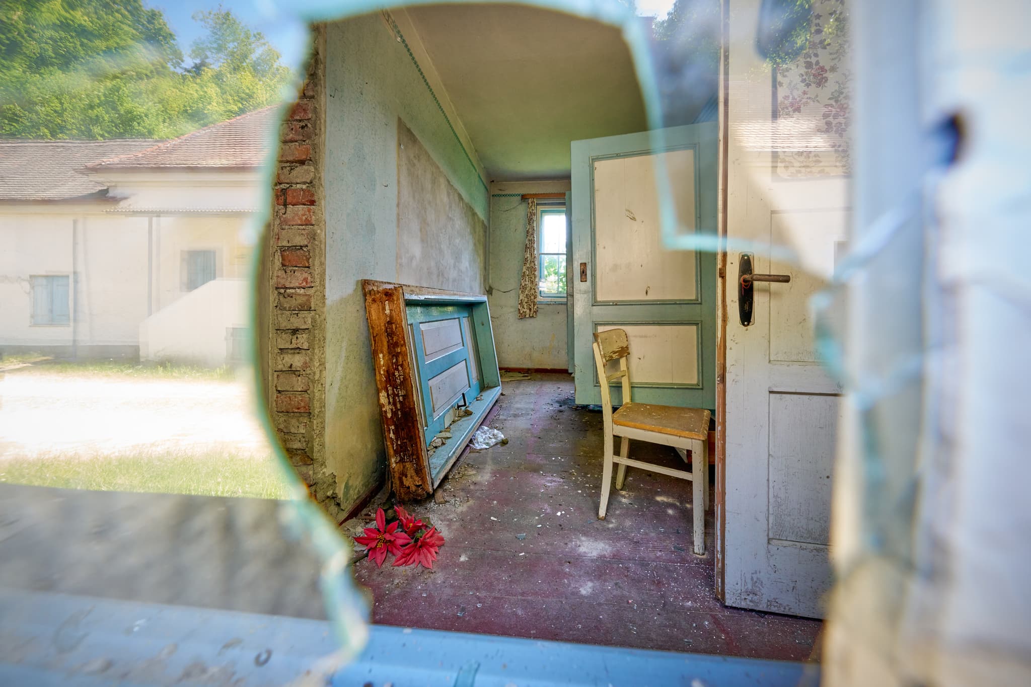 Blick durch zerbrochenes Fenster in eine alte Wäscherei in Ecksberg, Mühldorf. Das Innere zeigt verlassene Räume mit einem Stuhl und umgestürzten Möbeln.