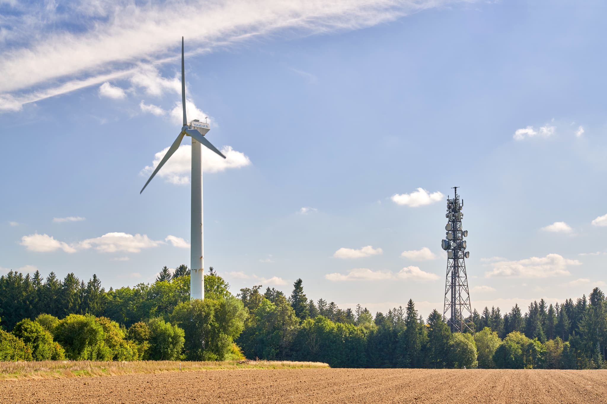 Windkraftanlage und Sendemast auf Feld vor Waldhügeln unter blauem Himmel mit Wolken. Standort Schnaitsee, Traunstein, Oberbayern, Chiemgau, Deutschland.