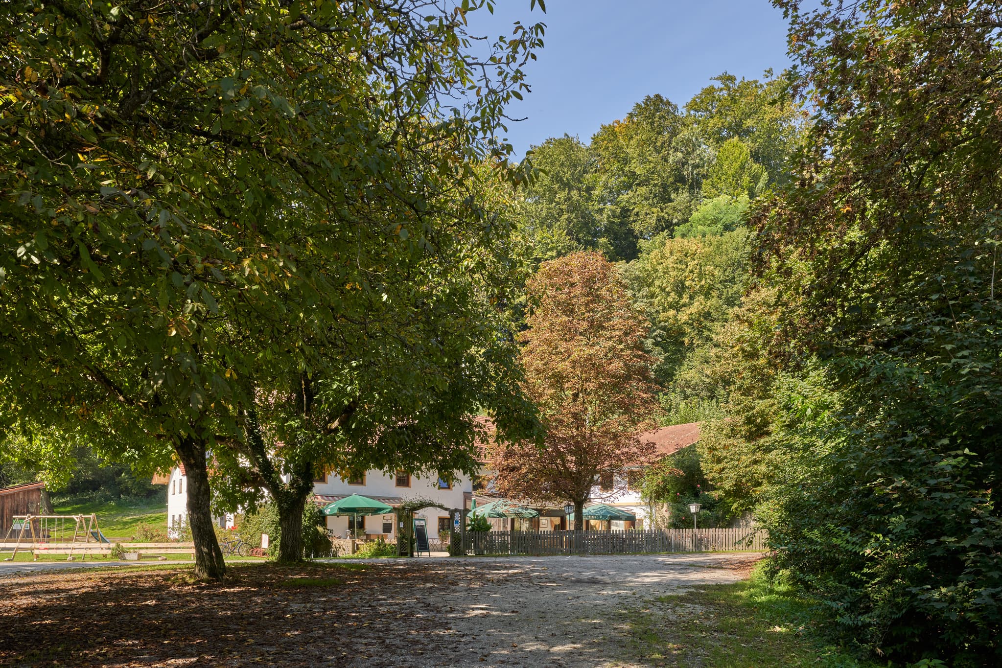 Gasthaus in Tiefenau, Burghausen, Landkreis Altötting, Oberbayern. Gebäude inmitten grüner Bäume, einen sonnigen Weg und einen Spielplatzbereich.