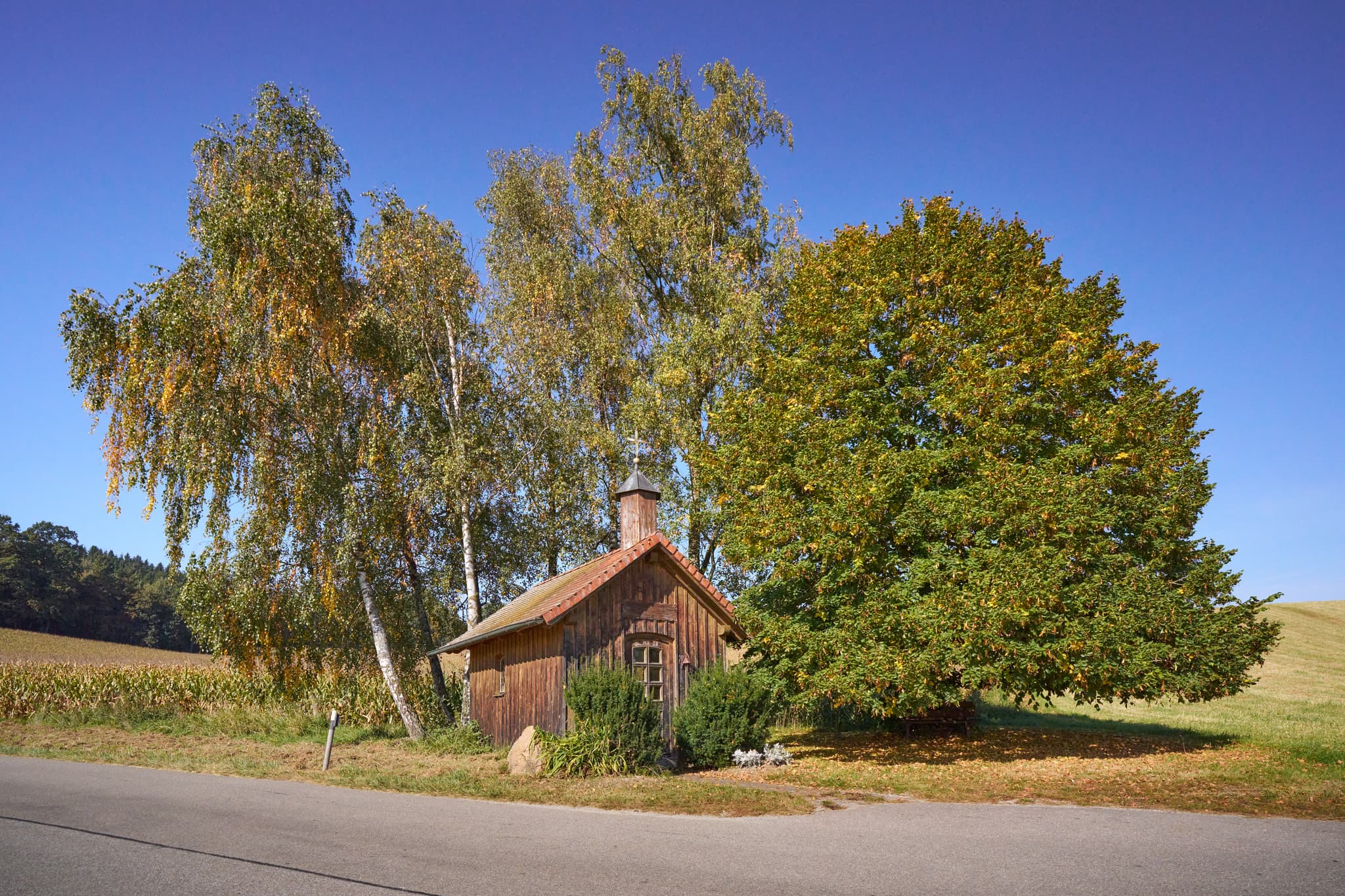 Holz-Kapelle in Stadlreith, Tettenweis im Landkreis Passau, Niederbayern, Deutschland. Umgeben von Bäumen und Feldern im Bayerischen Wald unter blauem Himmel.