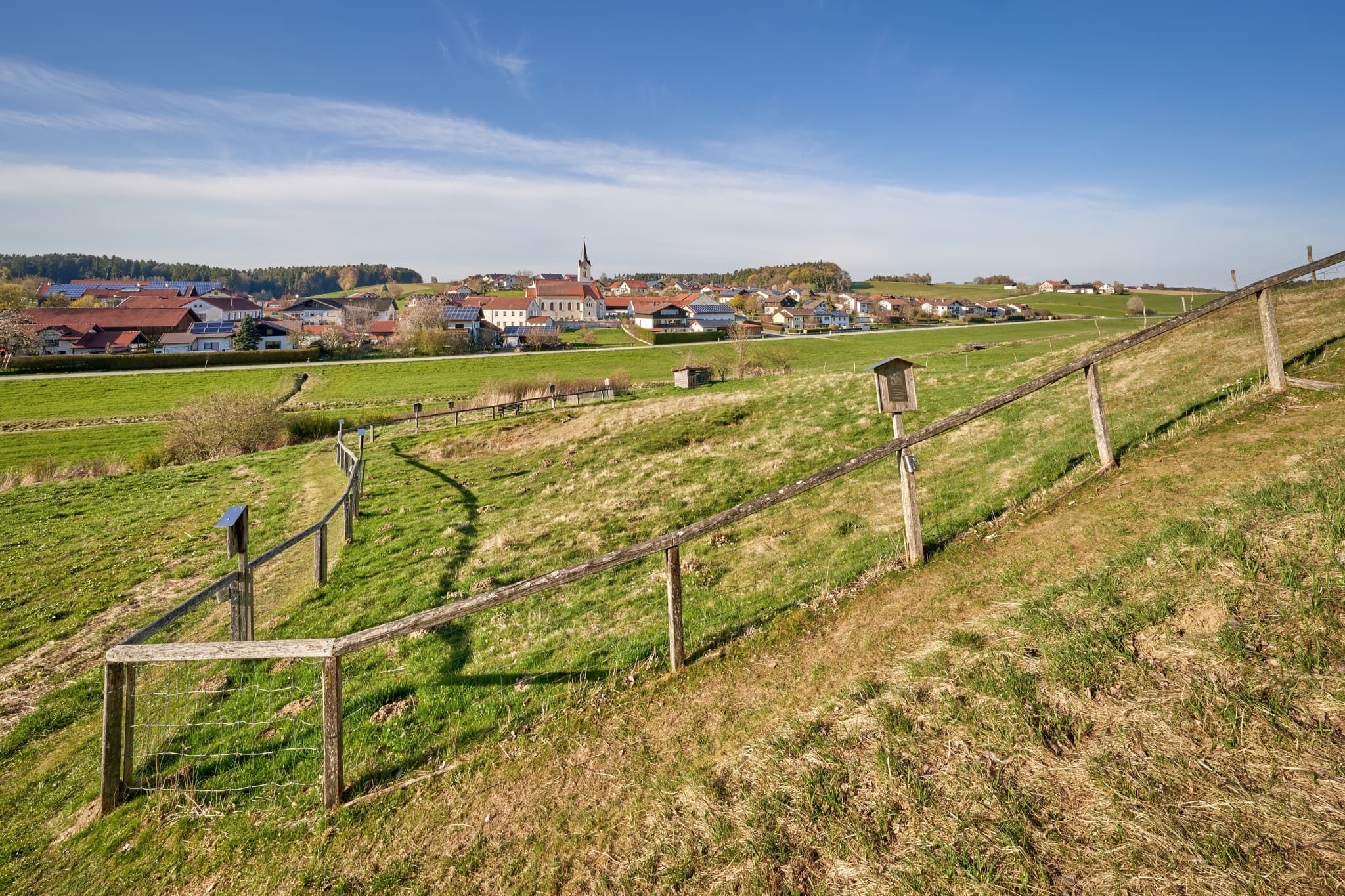 Landschaftsaufnahme am Kreuzweg in Erlbach im Landkreis Altötting, Oberbayern, Deutschland. Grüne Wiesen, ein Weg mit Zaun und das Dorf im Inn-Salzach-Gebiet