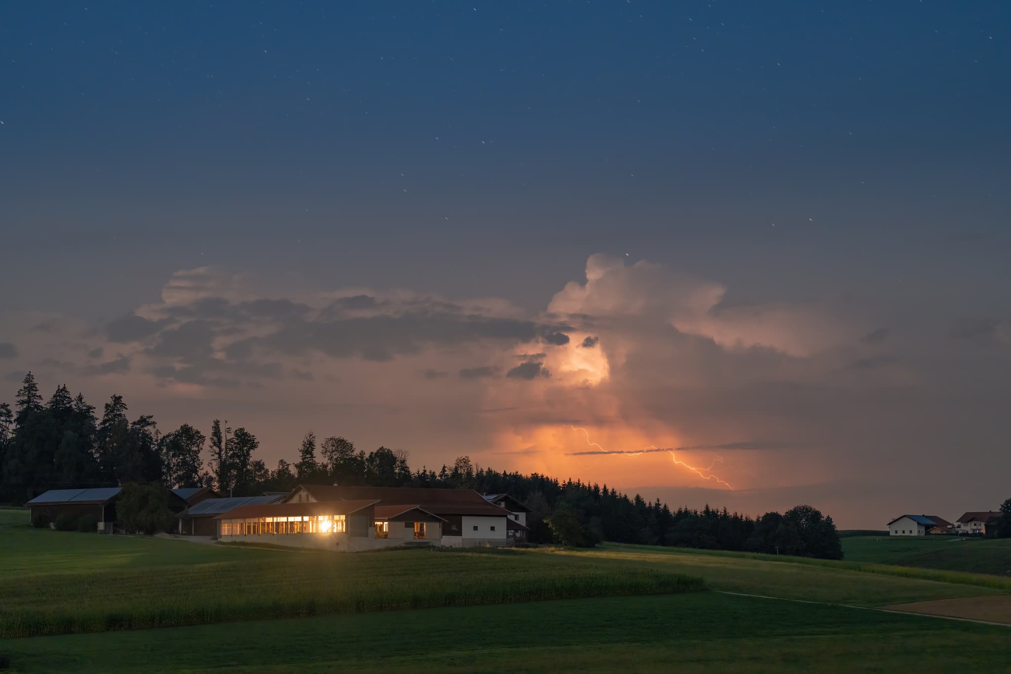 Gewittersturm über Reichwald, Reischach, Altötting, Oberbayern, Deutschland. Blitze erhellen Wolken über beleuchtetem Hof in der Inn-Salzach Region.