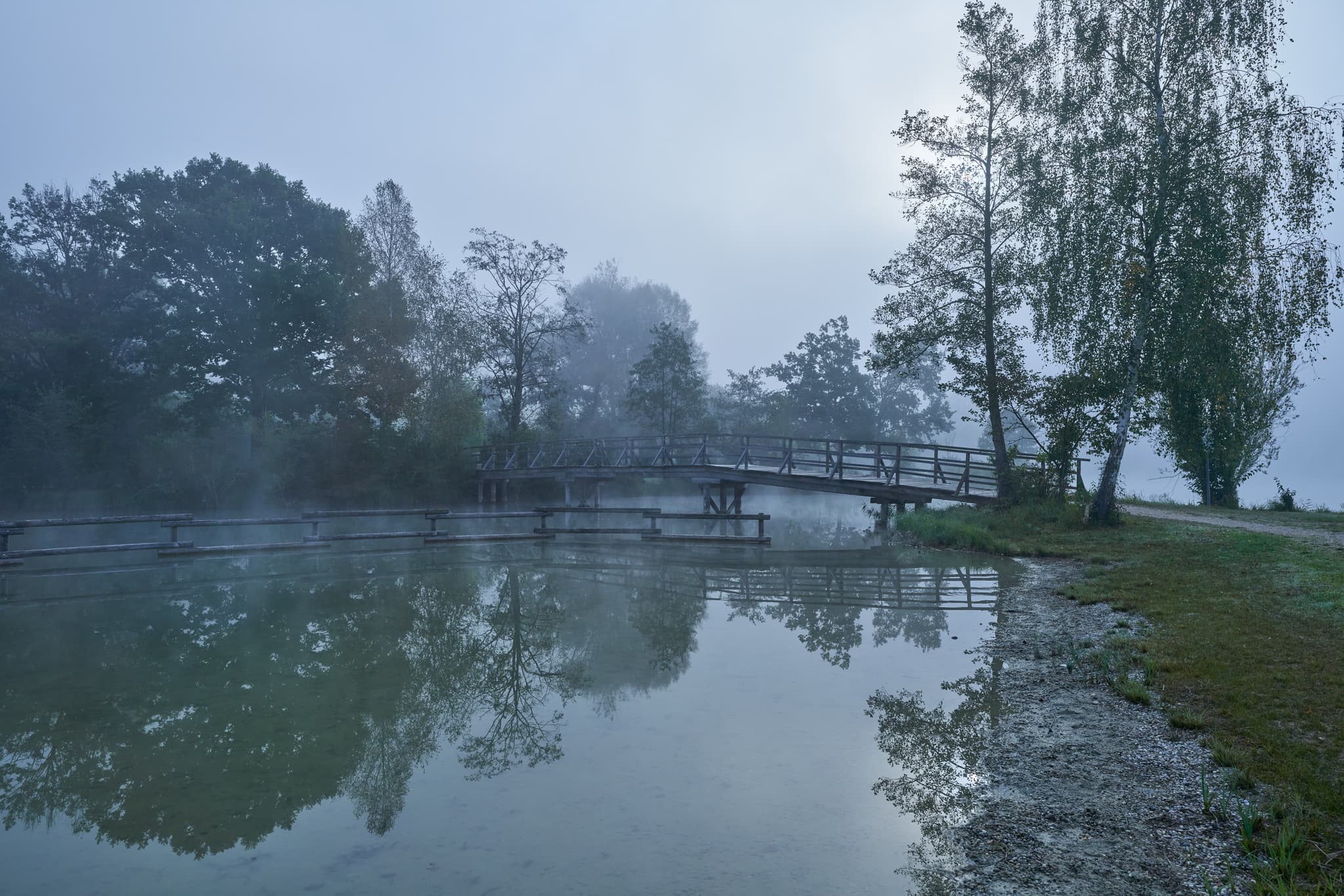 Badesee in Marktl, Landkreis Altötting, Oberbayern, Deutschland, im morgendlichen Herbstnebel. Die Inn-Salzach Landschaft mit See, Bäumen und Brücke.