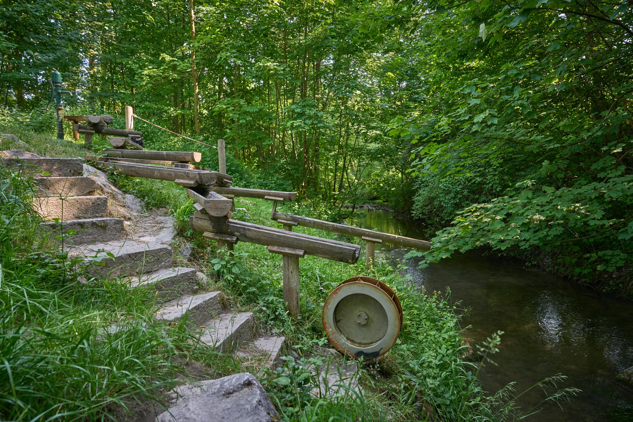 Idyllische Szene Naturerlebnisweg Innaue in Mühldorf am Inn, Oberbayern: Wasserspiel aus Holz am Bachlauf, Steinstufen in grüner Natur, Region Inn-Salzach.