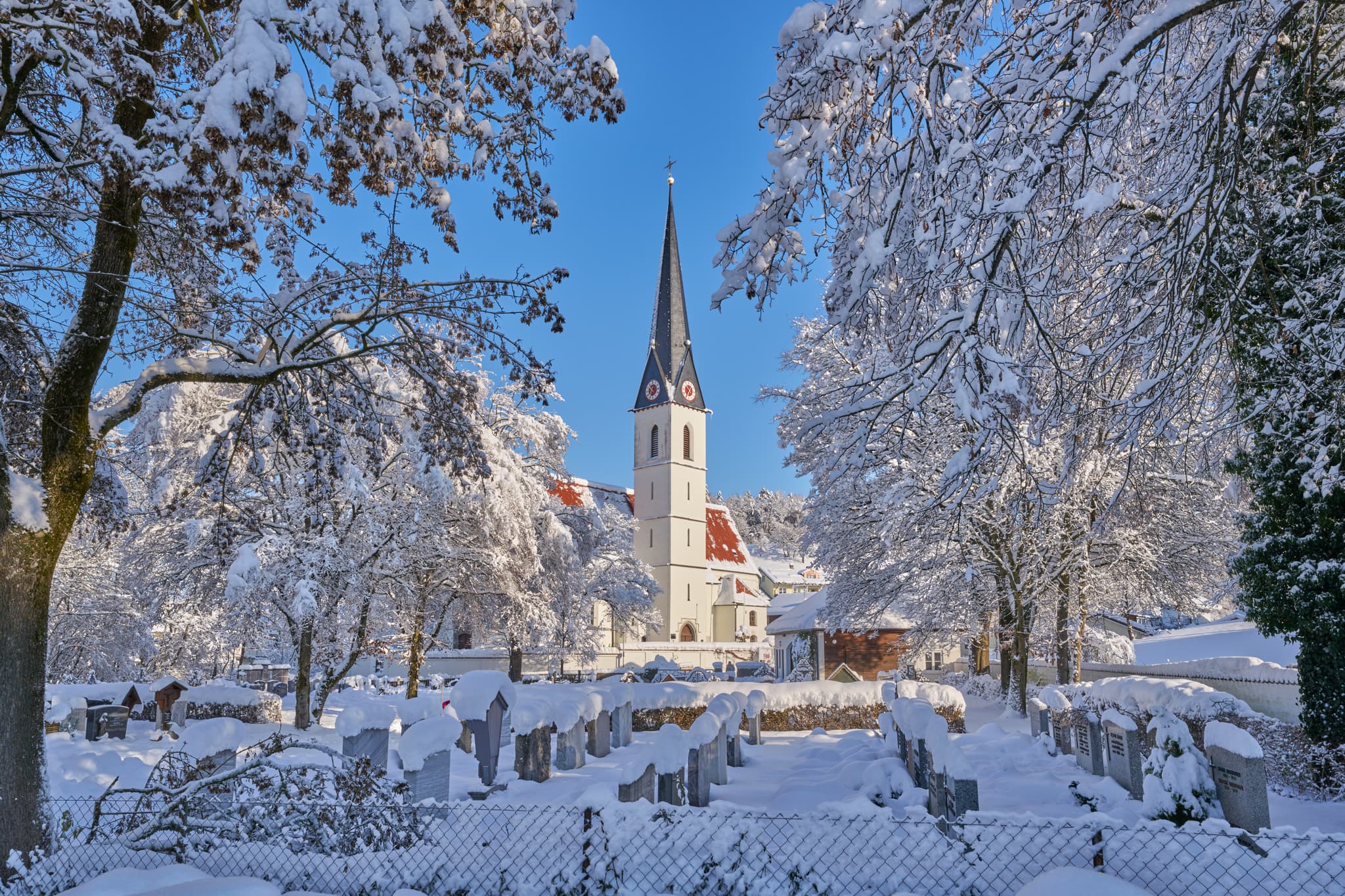 Pfarrkirche St. Martin Reischach, Altötting, Oberbayern, Deutschland, im Winter. Verschneite Kirche, Bäume, Friedhof. Blauer Himmel.