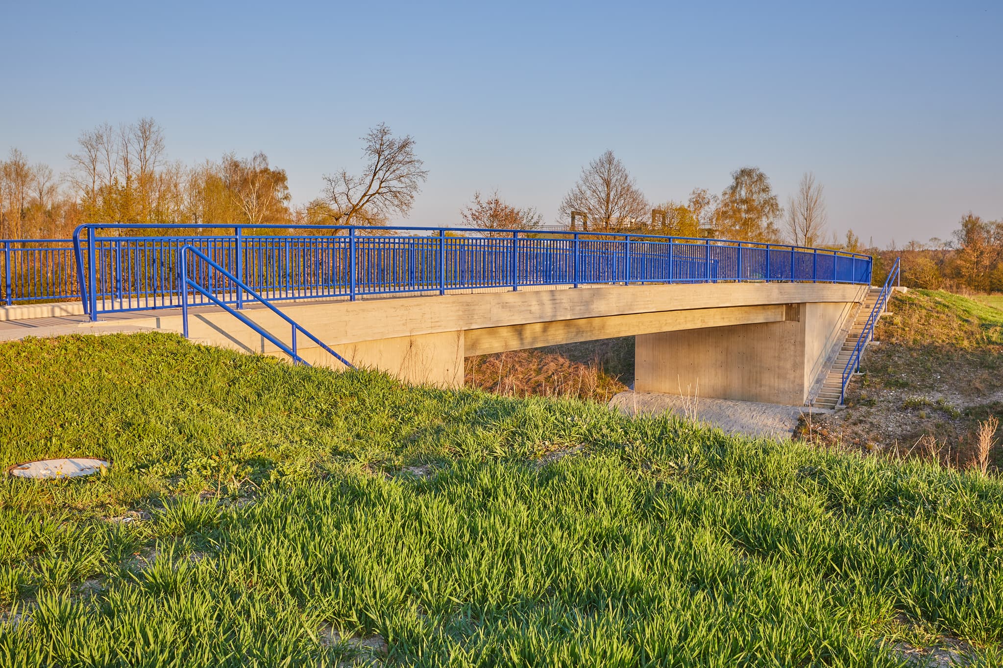 Moderne Brücke mit blauen Geländern am Innwerk Bahnübergang in Perach, Altötting, Oberbayern, Inn-Salzach, Deutschland, ist von Grünland umgeben.