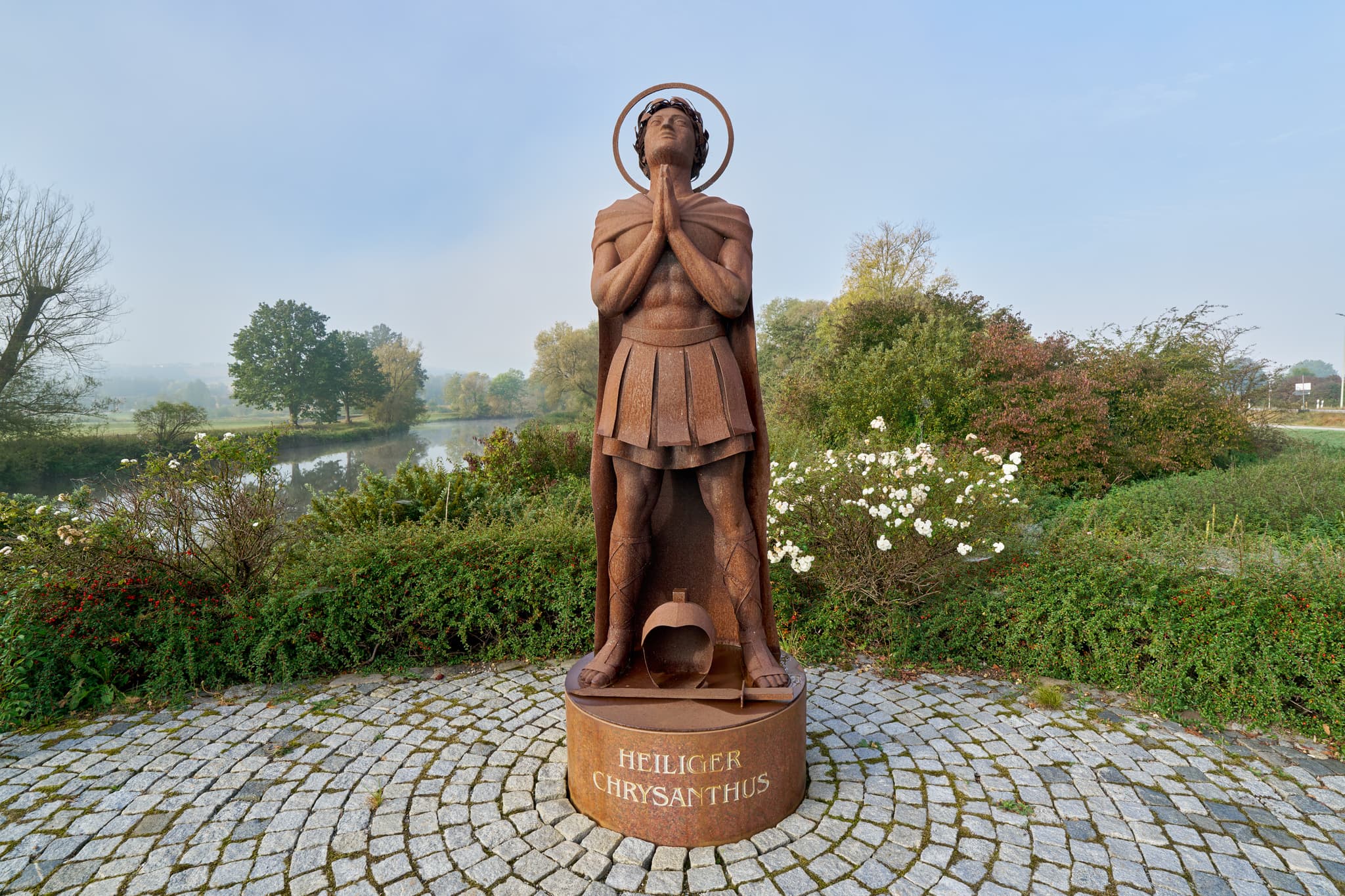Statue "Heiliger Chrysanthus" in Bad Birnbach, Rottal-Inn, Niederbayern. Herbstliche Szene am Ufer der Rott im Bäderdreieck, zeigt Figur auf Kopfsteinpflaster.