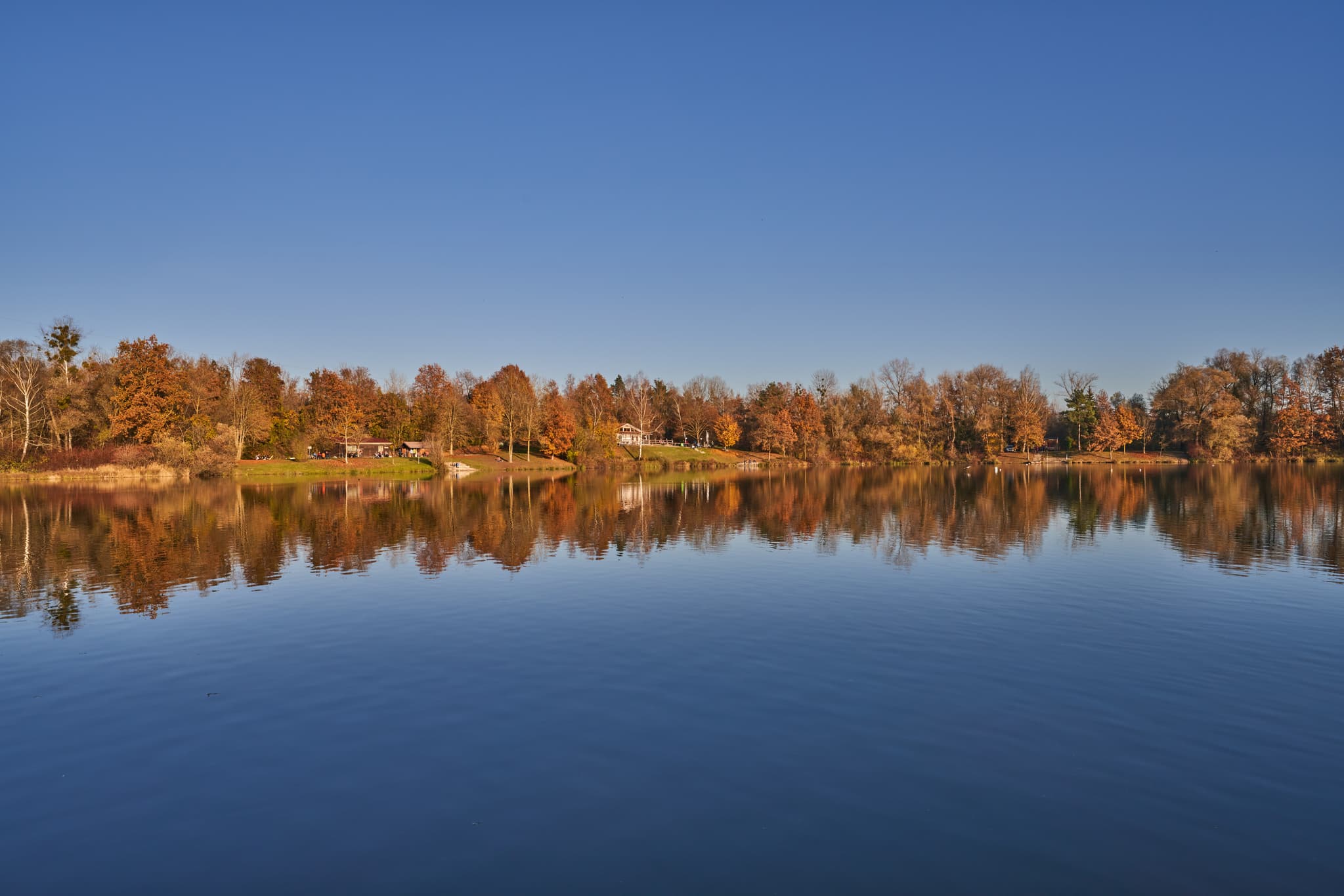 Herbstliche Naturaufnahme am Waldsee Lago bei Kirchdorf am Inn, Rottal-Inn, Niederbayern. Bunte Bäume spiegeln sich im ruhigen Wasser unter blauem Himmel.