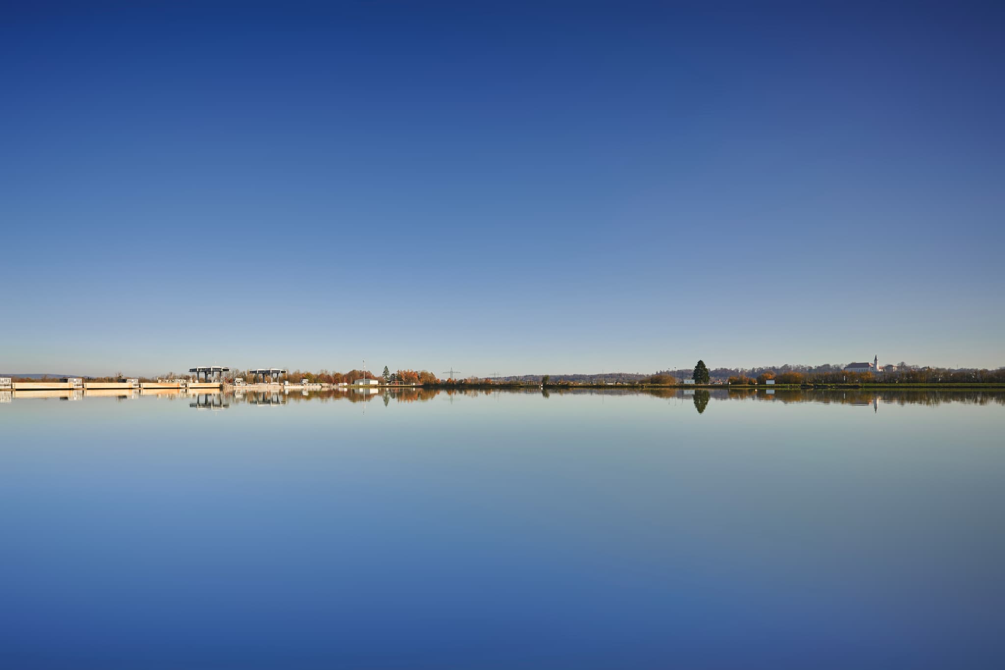 Das Kraftwerk Simbach-Braunau nahe Kirchdorf am Inn, Landkreis Rottal-Inn, Niederbayern. Weite Wasserfläche spiegelt blauen Himmel und Ufer.