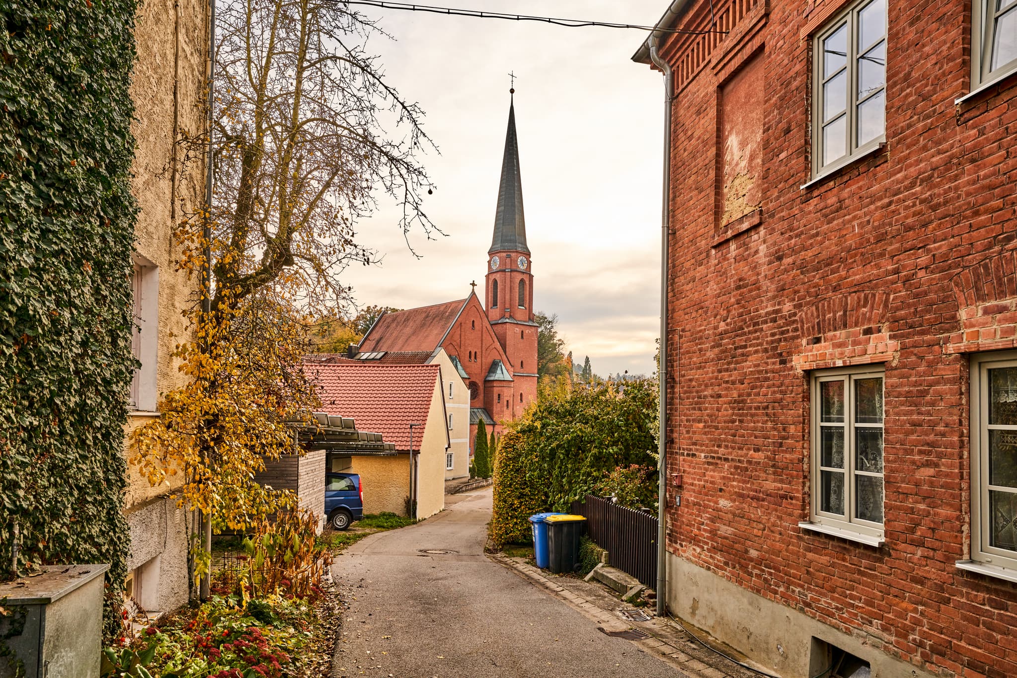 Blick von der Alte Schule Hörniweg in Schönau, Landkreis Rottal-Inn, Niederbayern, Deutschland. Gasse mit Backsteinhaus und  Kirche im herbstlichen Holzland.