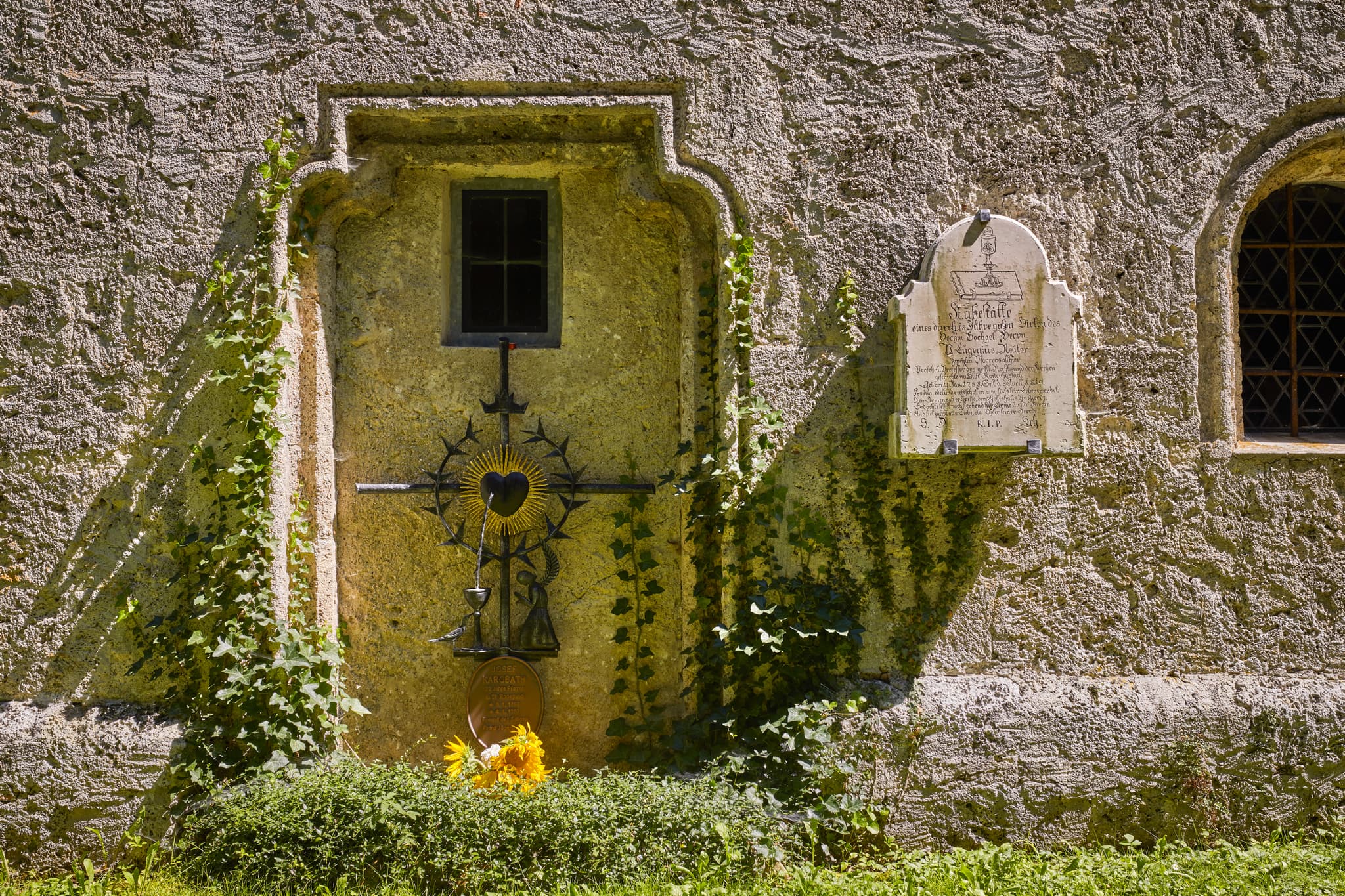 Kirchenmauer mit kunstvollem Metallkreuz und Steintafel. Aufnahme aus St. Radegund, Landkreis Braunau am Inn, Innviertel, Oberösterreich, Österreich.
