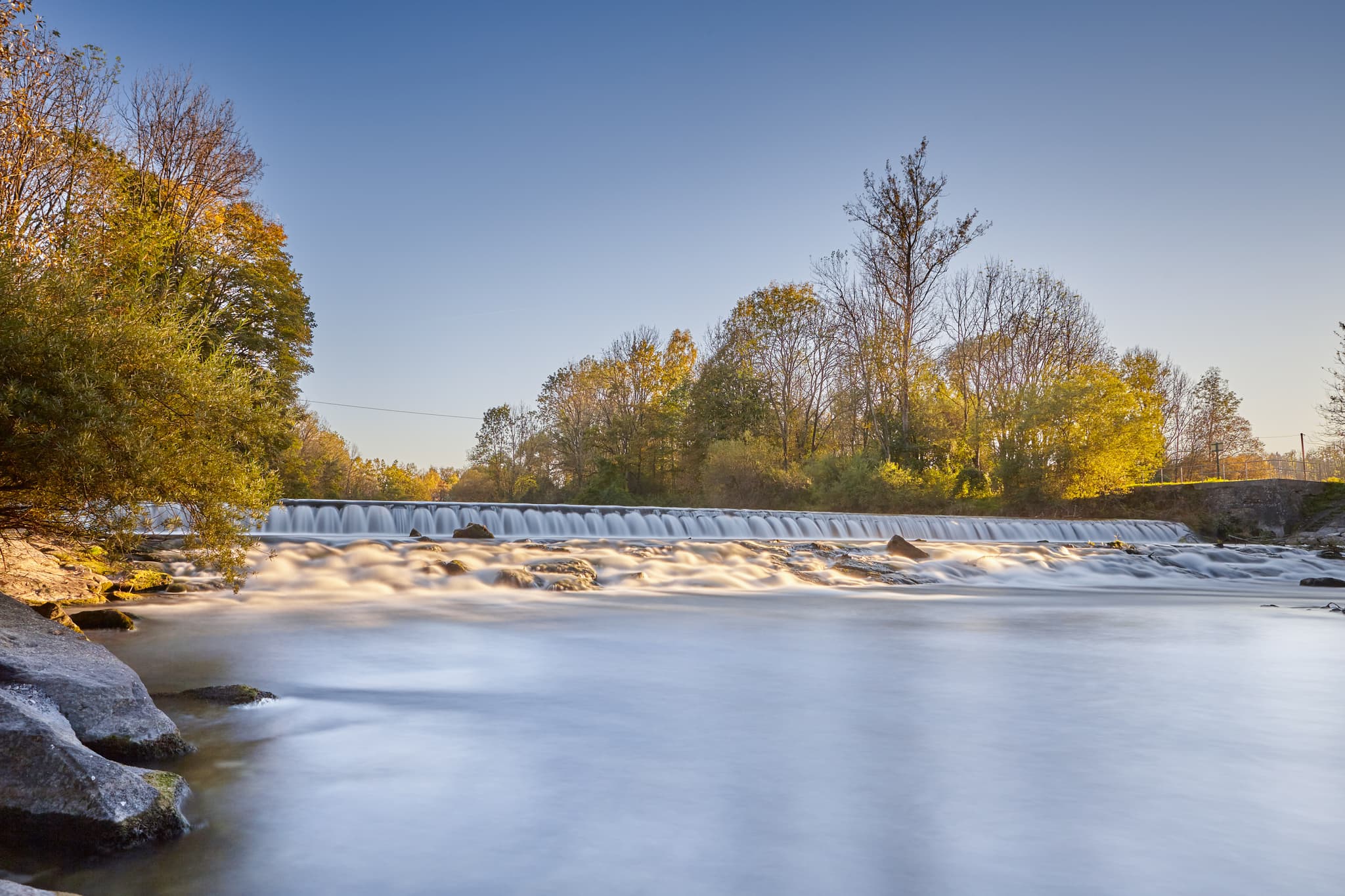 Obere Alz, Wald, Garching, Landkreis Altötting, Oberbayern. Eine Flusslandschaft der Inn-Salzach Region, Deutschland, mit Wasserfall, Bäumen und Herbstfarben.