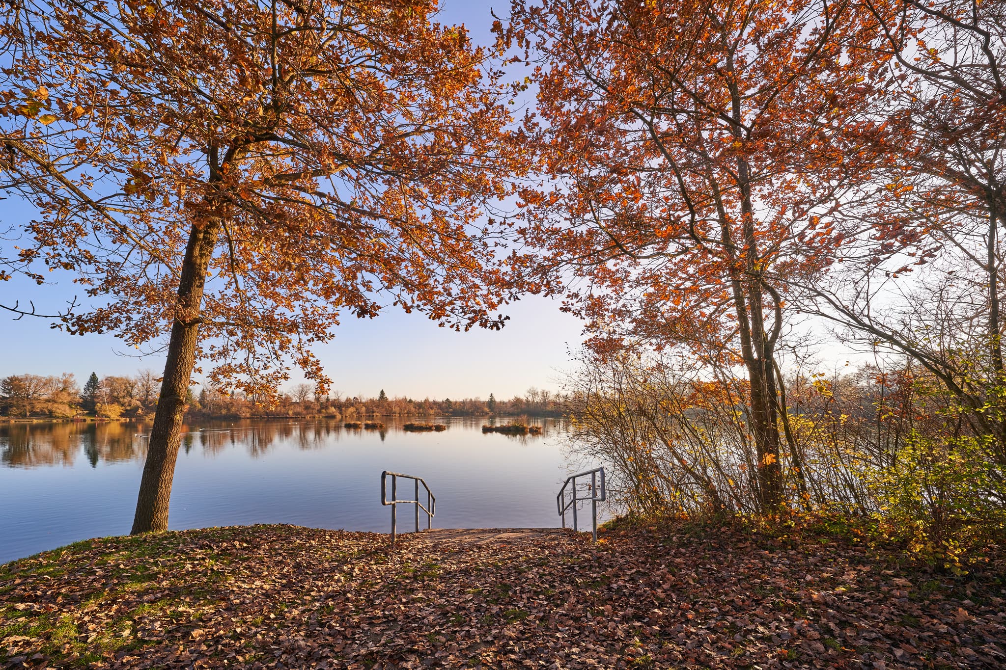 Herbstlicher Waldsee, Kirchdorf am Inn, Rottal-Inn, Niederbayern, Deutschland. Ruhiger See, bunte Bäume, Herbstlaub am Ufer. Treppe ins Wasser.