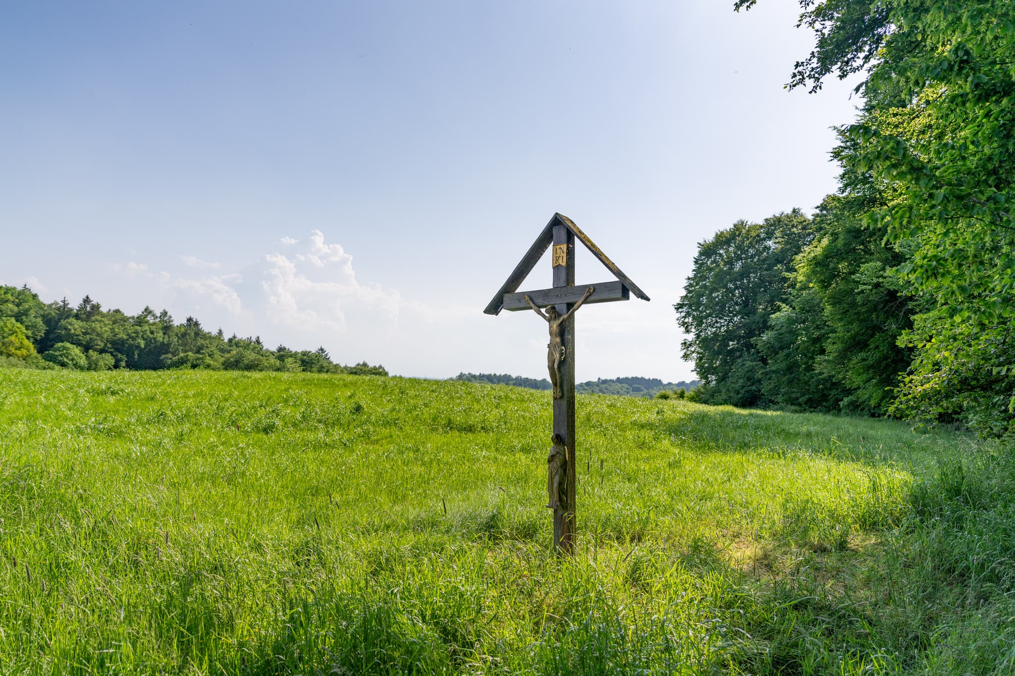 Ein Wegkreuz steht in einem weiten grünen Feld nahe Friesing, einer Ortschaft in Reischach, Landkreis Altötting, Oberbayern, Inn-Salzach, Deutschland.
