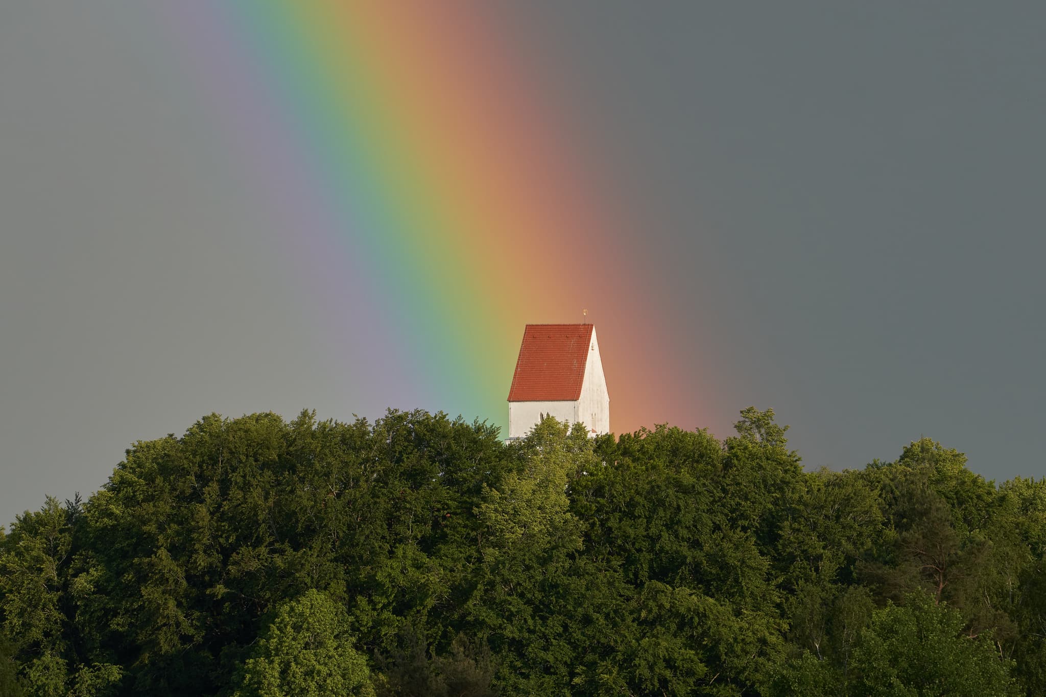 Ein farbenprächtiger Regenbogen spannt sich über einem bewaldeten Hügel bei Steinhausen, Gemeinde Erlbach. Die Landschaft im Landkreis Altötting, Oberbayern.