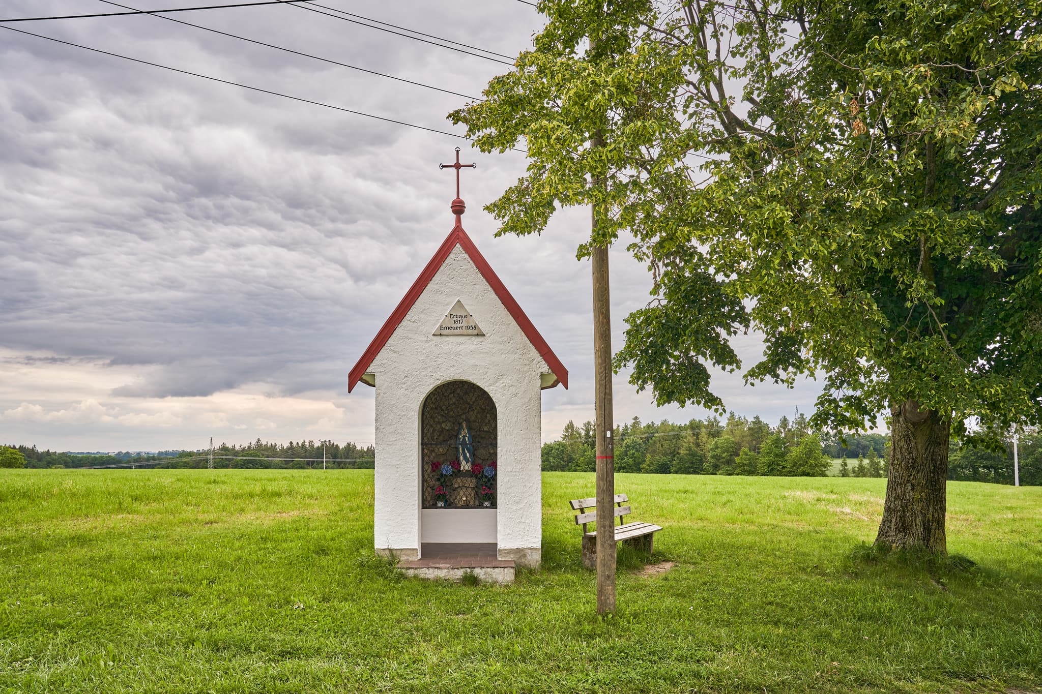 Kapelle Höresham, Burgkirchen, Altötting, Oberbayern. In grüner Wiesenlandschaft Inn-Salzach, Deutschland. Baum & Bank unter bewölktem Himmel.