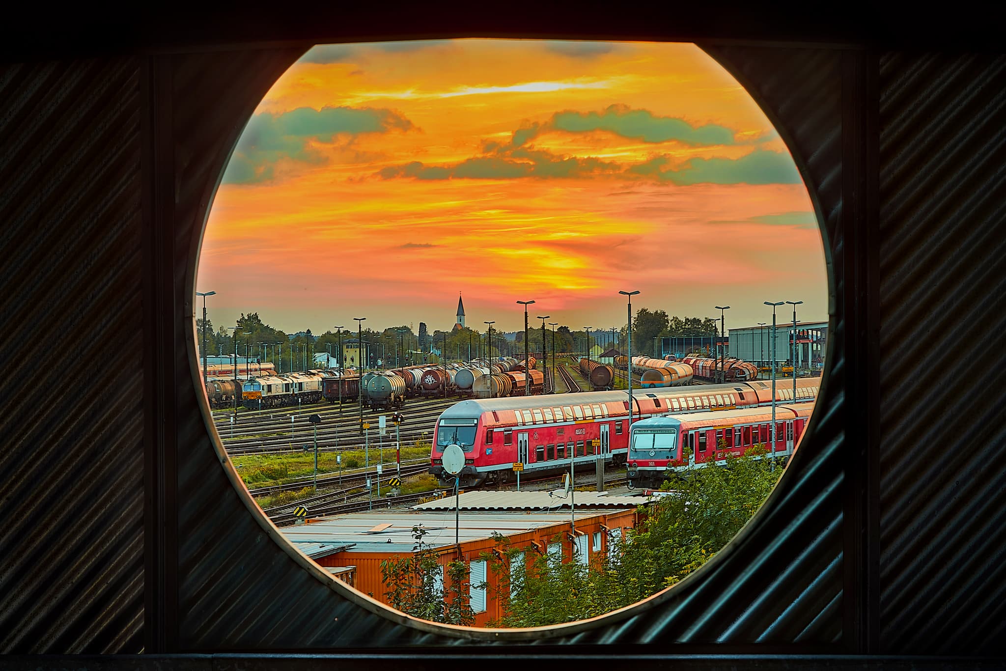 Blick durch ein Fenster auf den Bahnhof Mühldorf am Inn. Zuggleise und Gebäude des Paarkhauses unter Abendrot.