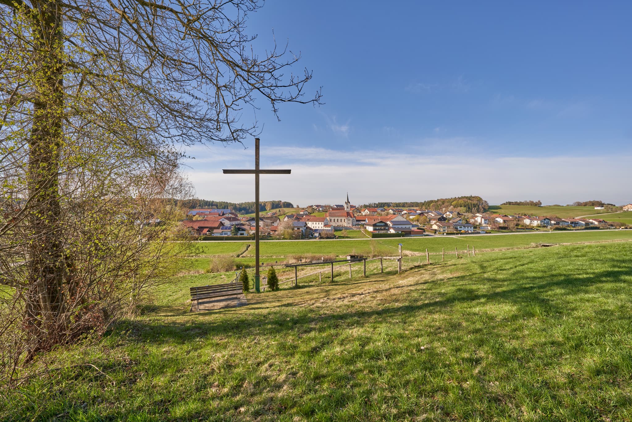 Ansicht Kreuzweg in Erlbach, Altötting, Oberbayern. Holzkreuz mit Bank vor Dorf. Grüne Wiesen, blauer Himmel. Region Inn-Salzach, Deutschland.