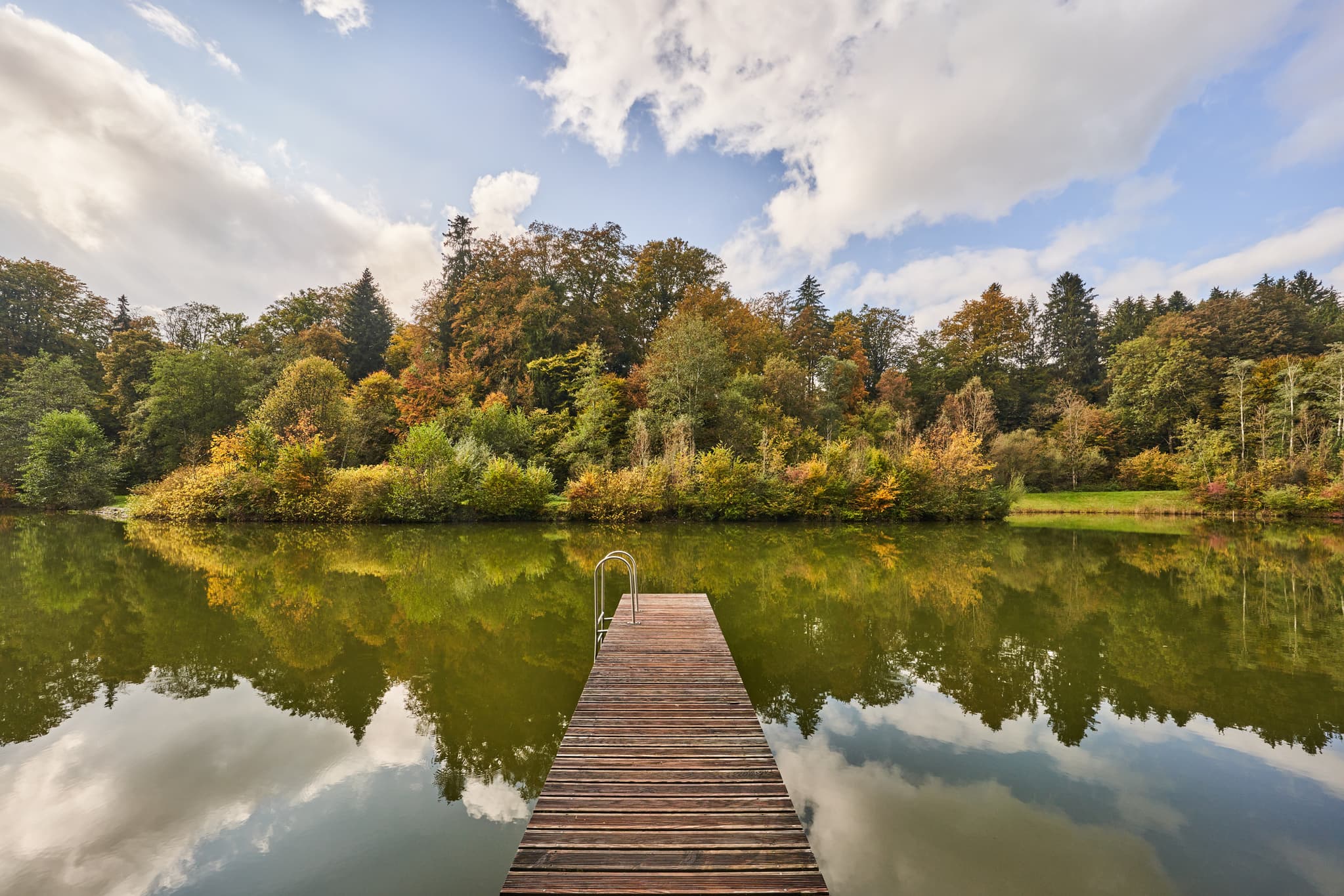Herbst am Hochmühl Badesee bei Reischach im Landkreis Altötting, Oberbayern, Region Inn-Salzach, Deutschland. Ruhiger See, Holzsteg, farbenfroher Baumbewuchs.