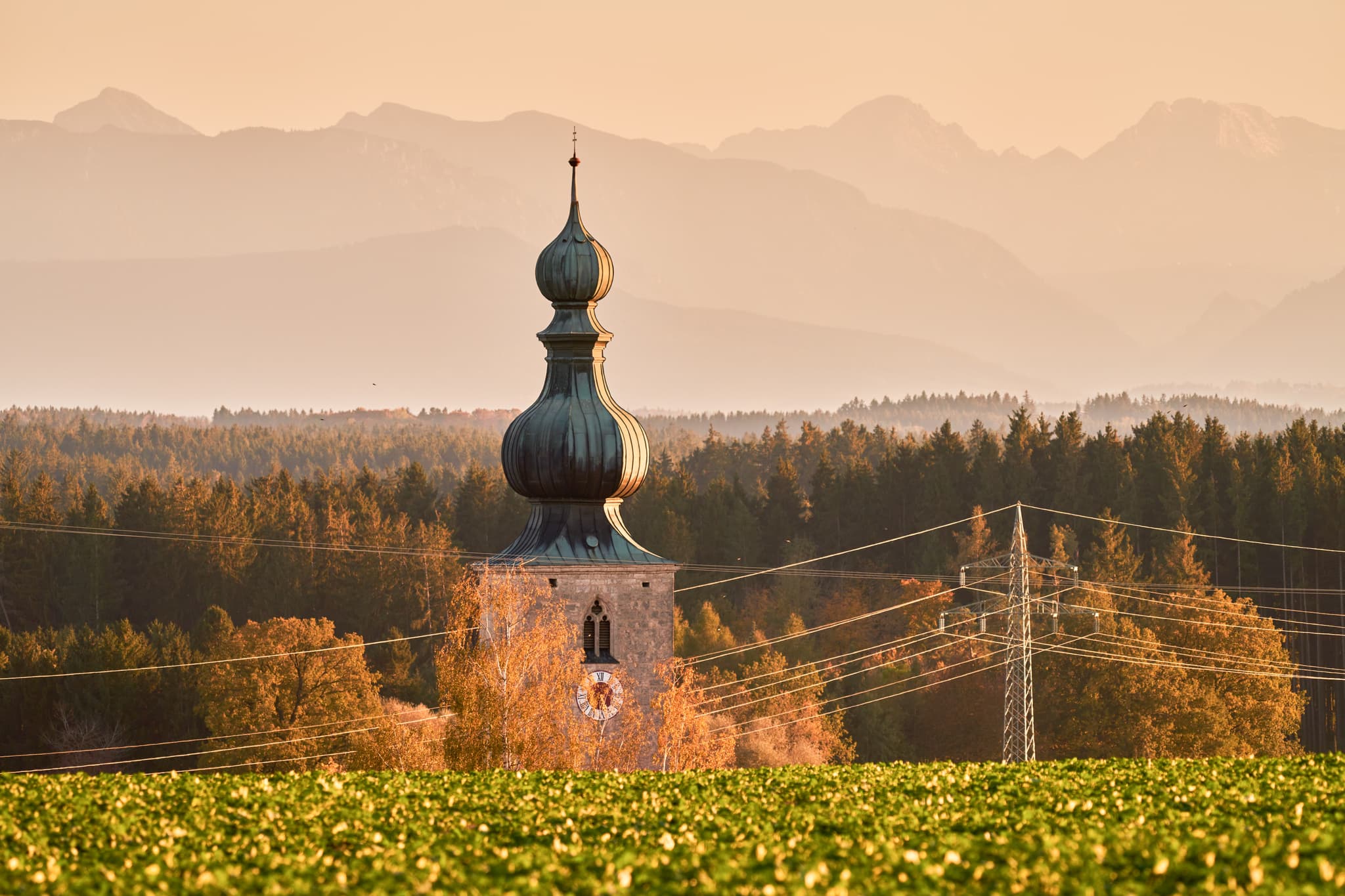 Abendliche Szene mit Zwiebelturm-Kirche in Rainbichl, Tyrlaching, Landkreis Altötting, Oberbayern, Inn-Salzach, Deutschland. Felder, Wälder, Berge, Strommast.