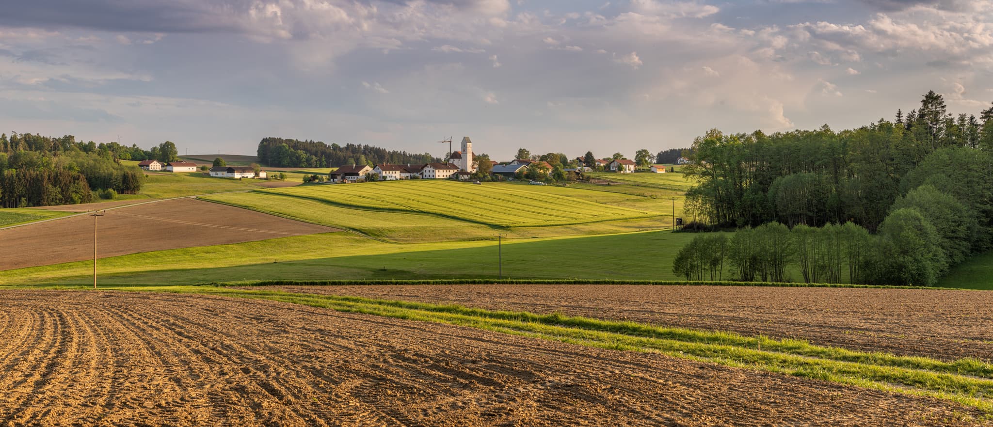 Ländliche Landschaft bei Ecking, Reischach, Altötting, Oberbayern, Inn-Salzach, Deutschland. Felder, Hügel und ein Dorf mit Kirche unter bewölktem Himmel.