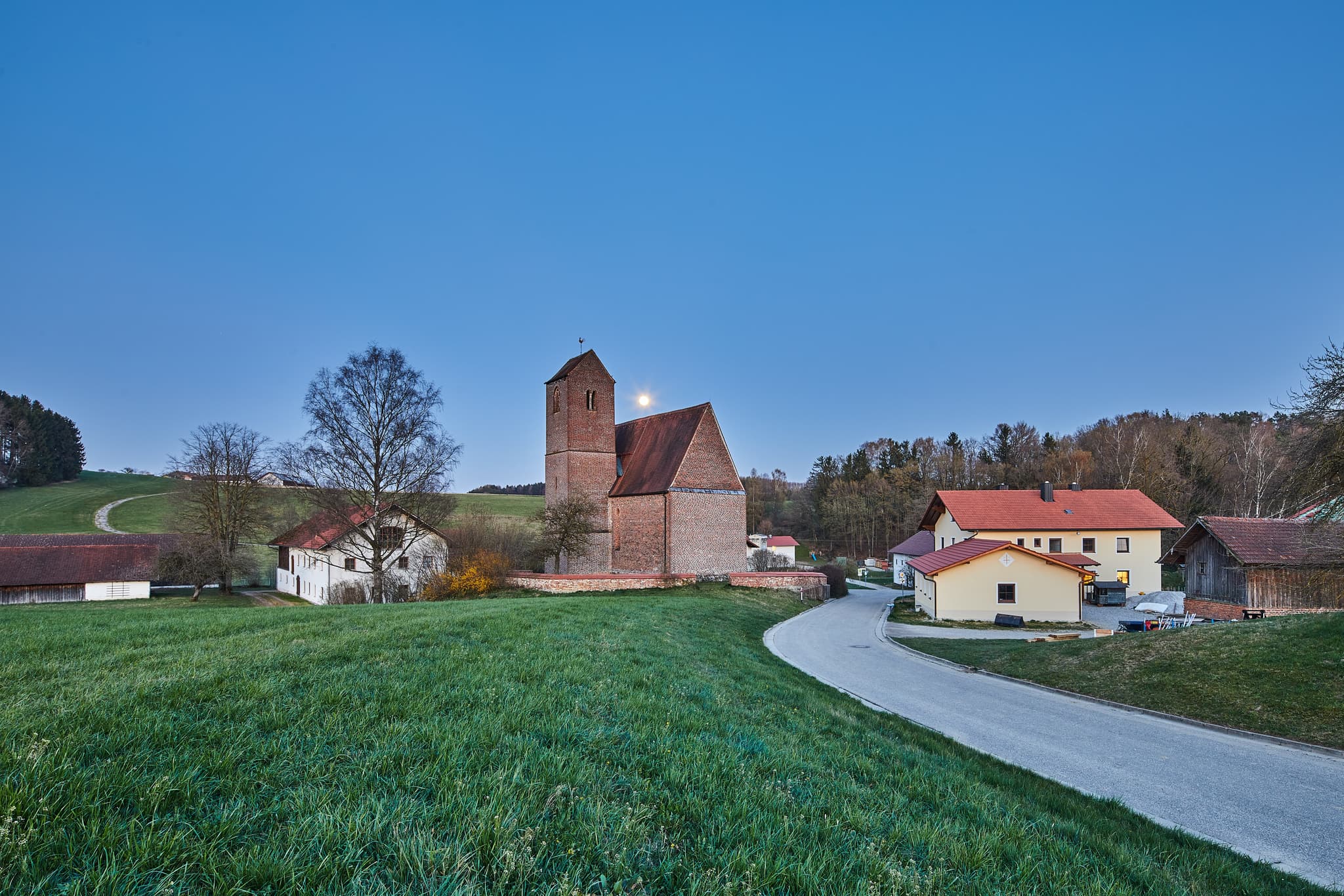 Ländliche Kirche in Gehersdorf, Zeilarn, Rottal-Inn, Niederbayern, Deutschland. Blaue Stunde mit Mond über sanften Hügeln und grünen Feldern im Holzland.