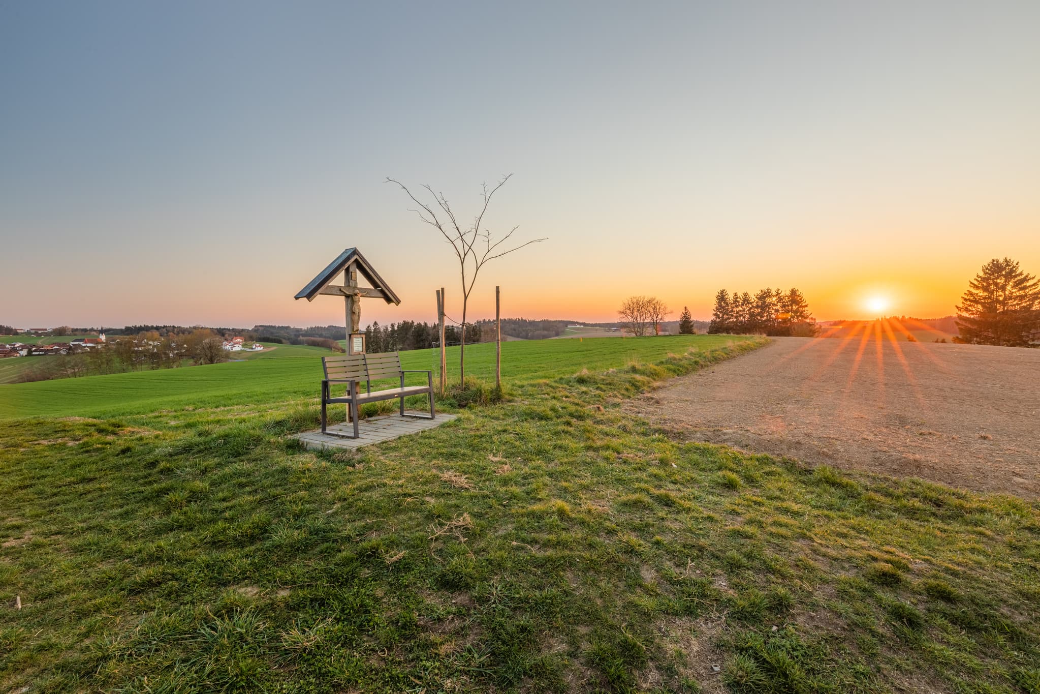 Abendsonne über einem Feldkreuz mit Bank bei Arbing in der Gemeinde Reischach, Landkreis Altötting, Oberbayern, Bayern, Deutschland