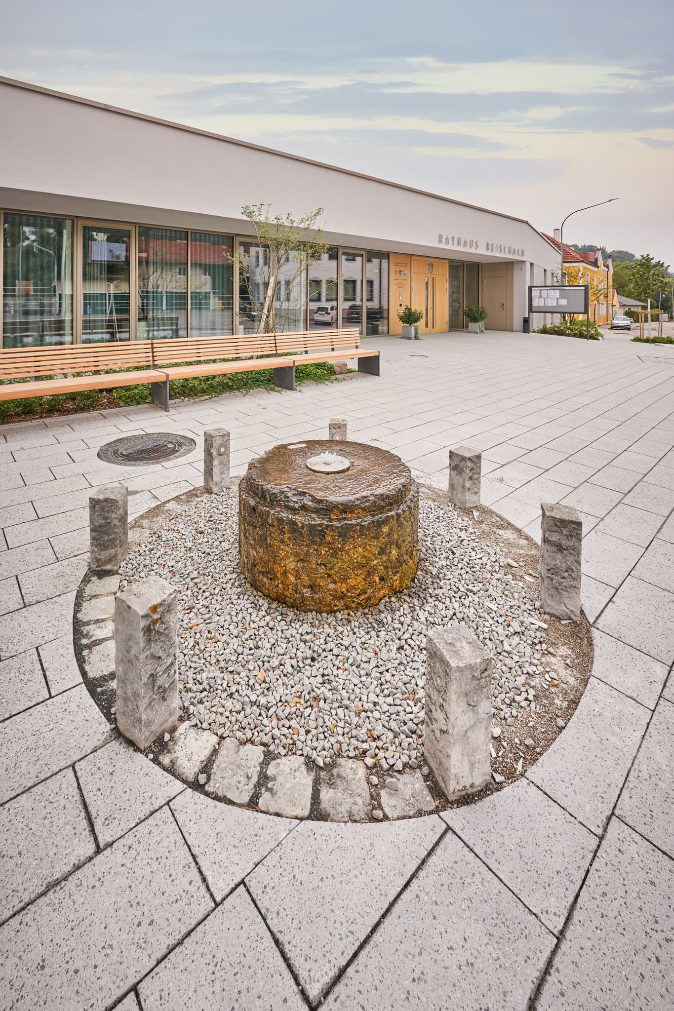 Ein historischer Mühlsteinbrunnen steht zentral auf einem gepflasterten Platz in Gemeinde Brunnen, Reischach, Altötting, Inn-Salzach in Oberbayern, Deutschland.