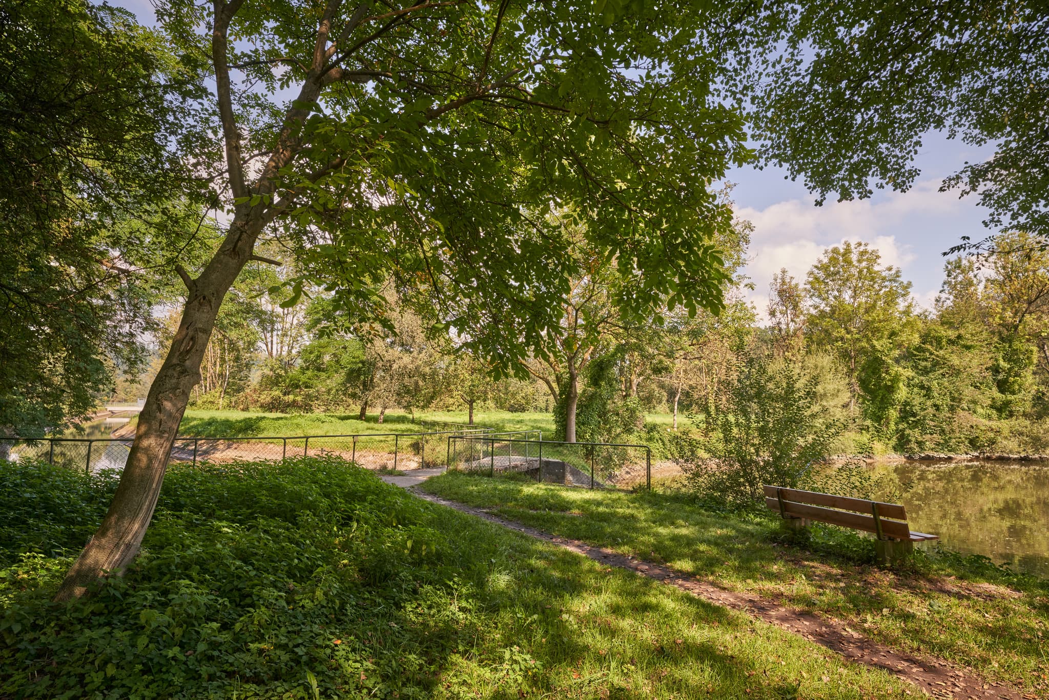 Landschaft am Stauweiher bei Winhöring, Landkreis Altötting, Oberbayern. Grünes Ufer mit Bäumen, Weg und Sitzbank am Wasser. Region Inn-Salzach, Deutschland.