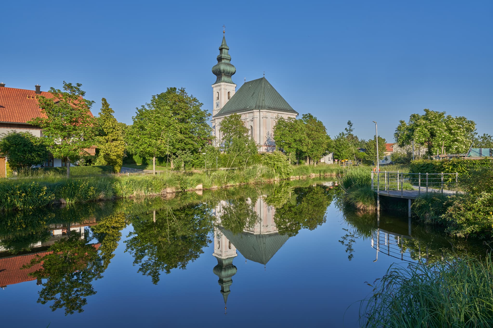 Die Dorfkirche von Kirchweidach spiegelt sich im Dorfweiher. Das Bild zeigt die Kirche, den Weiher und die umliegende Landschaft im Landkreis Altötting.