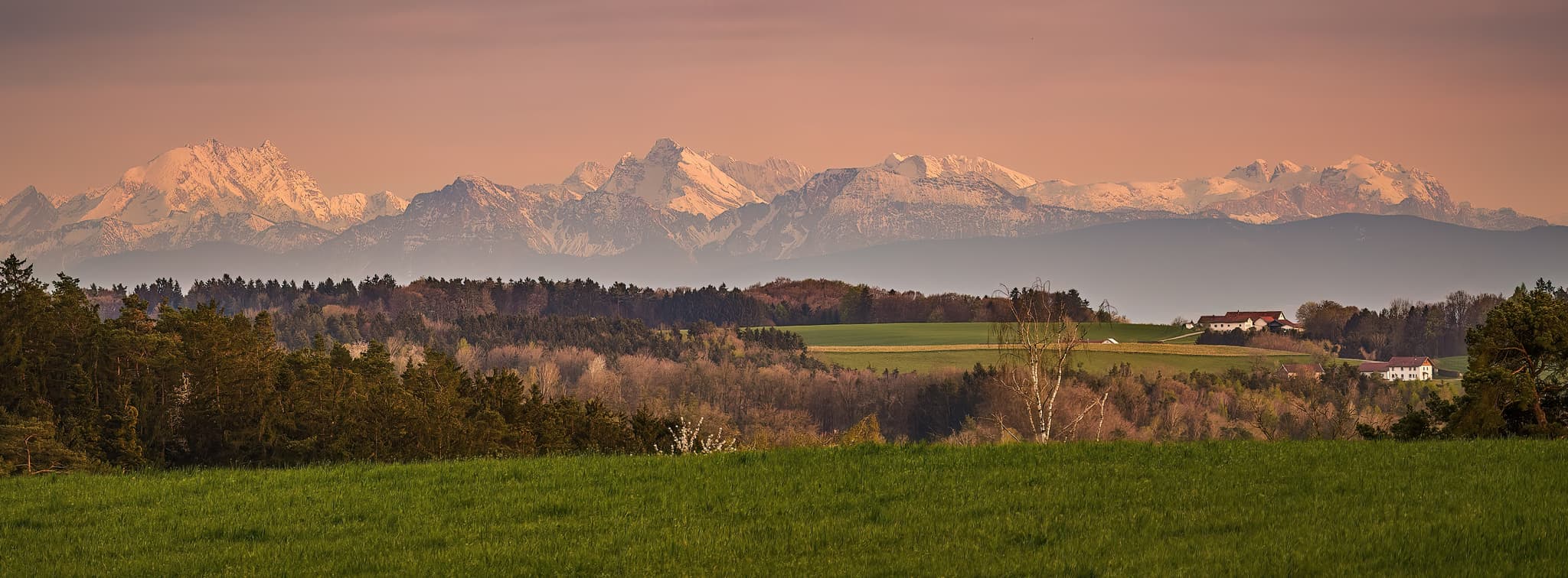 Aussicht von Waldberg, Reischach, Altötting, Oberbayern, Deutschland. Grüne Felder, Wälder und ferne Gebäude von Aushofen mit den Alpen im Hintergrund.