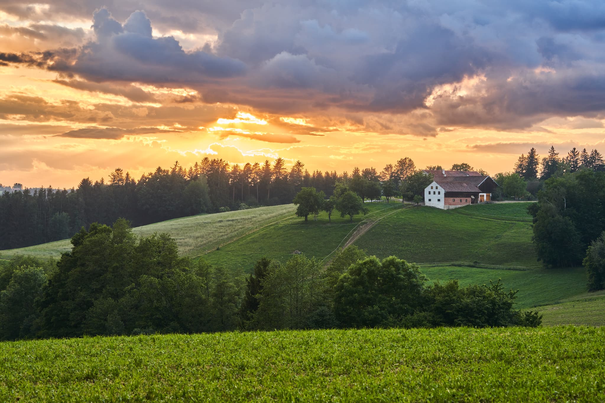Landschaftsbild Kienberg, Reischach, Altötting. Hof auf grünen Hügeln, Felder und Wälder unter dramatischem Gewitterhimmel mit Goldlicht.
