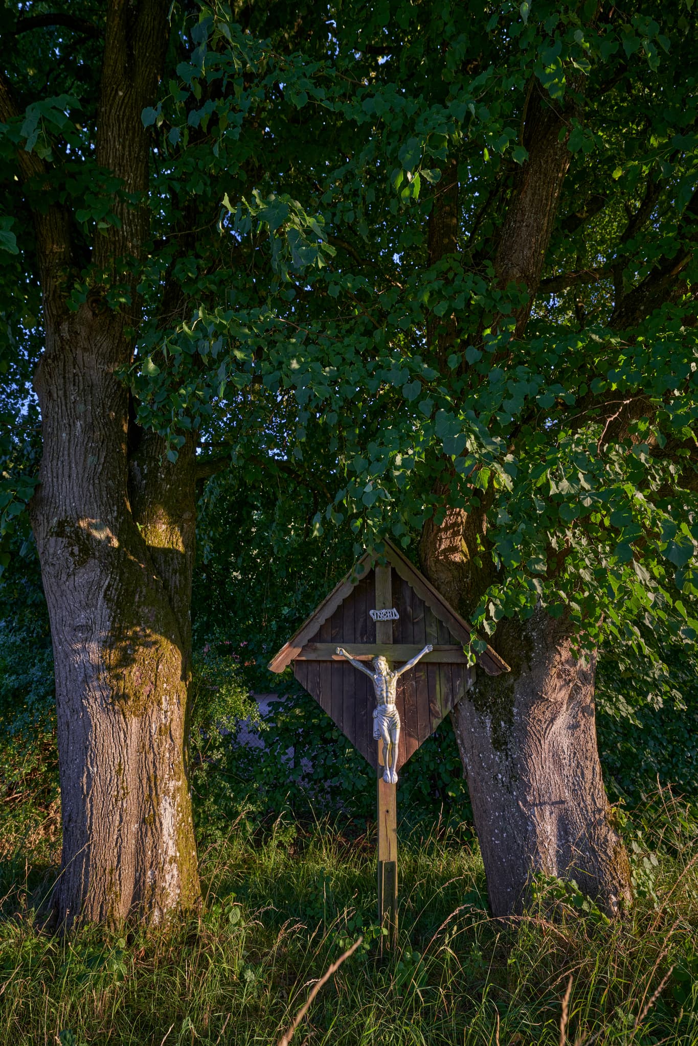 Wegkreuz am Wertstoffhof in Erlbach, Landkreis Altötting, Oberbayern. Das Motiv zeigt das Kreuz unter zwei großen Bäumen in der Region Inn-Salzach, Deutschland.