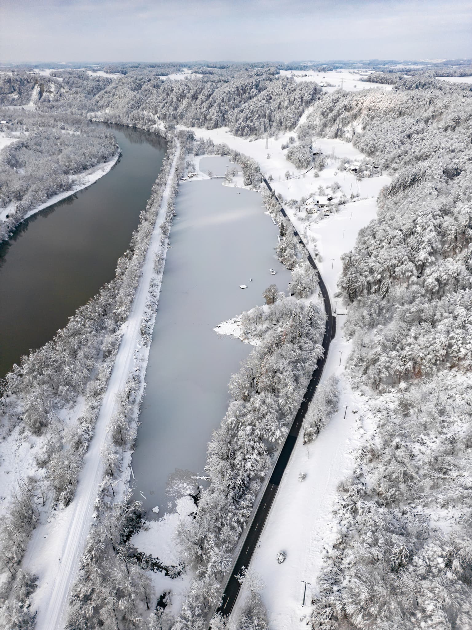 Badesee in Marktl, Landkreis Altötting, Oberbayern, Inn-Salzach, Deutschland. Die winterliche Landschaft zeigt See, Straße und schneebedeckte Umgebung.