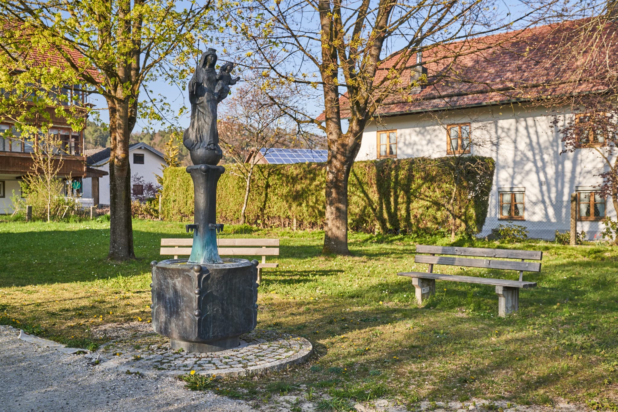 Brunnen mit Marienfigur und Sitzbänken auf Grünfläche vor der Kirche in Perach, Altötting, Inn-Salzach, Oberbayern, Deutschland.