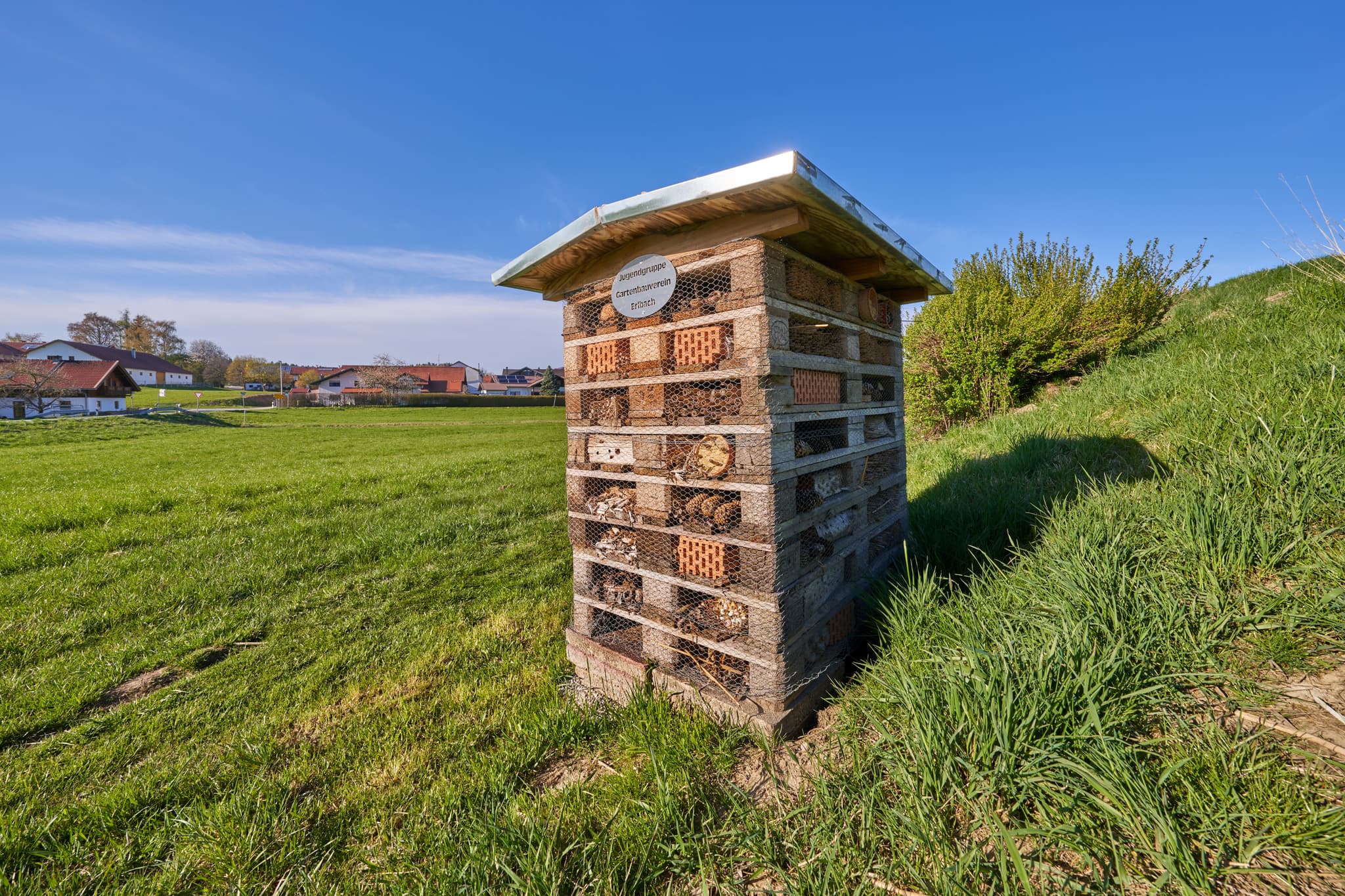 Insektenhotel am Kreuzweg in Erlbach, Landkreis Altötting, Oberbayern. Die Region Inn-Salzach in Deutschland zeigt weite Grünflächen unter blauem Himmel.