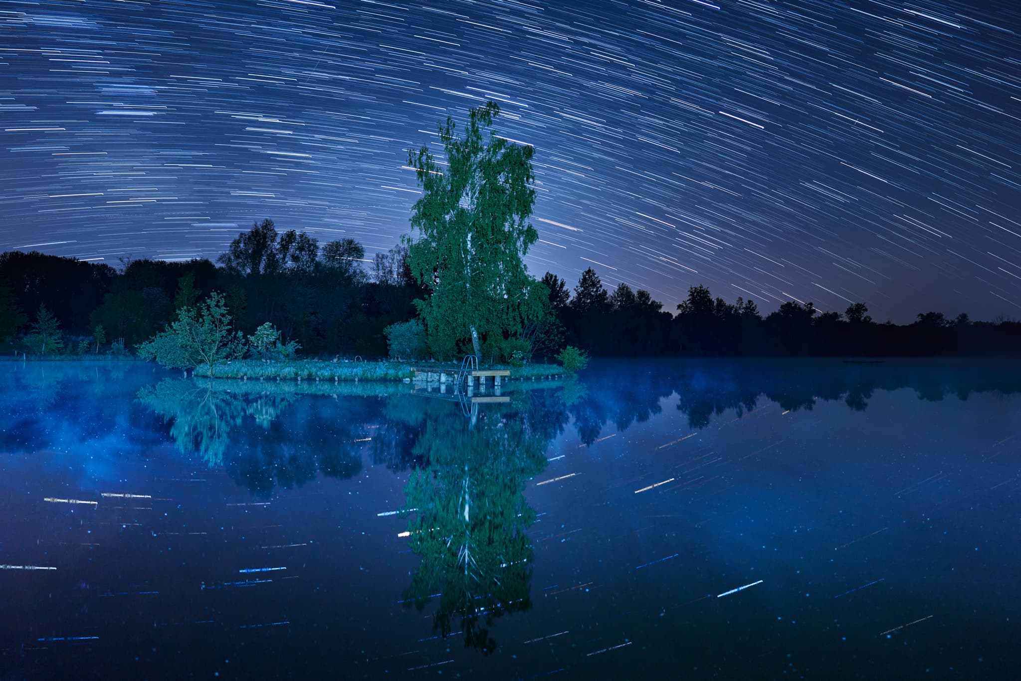 Nachtaufnahme eines Startrails über dem Peracher Badesee in Perach, Altötting, Oberbayern, Deutschland. Spiegelungen im See der Inn-Salzach Region.