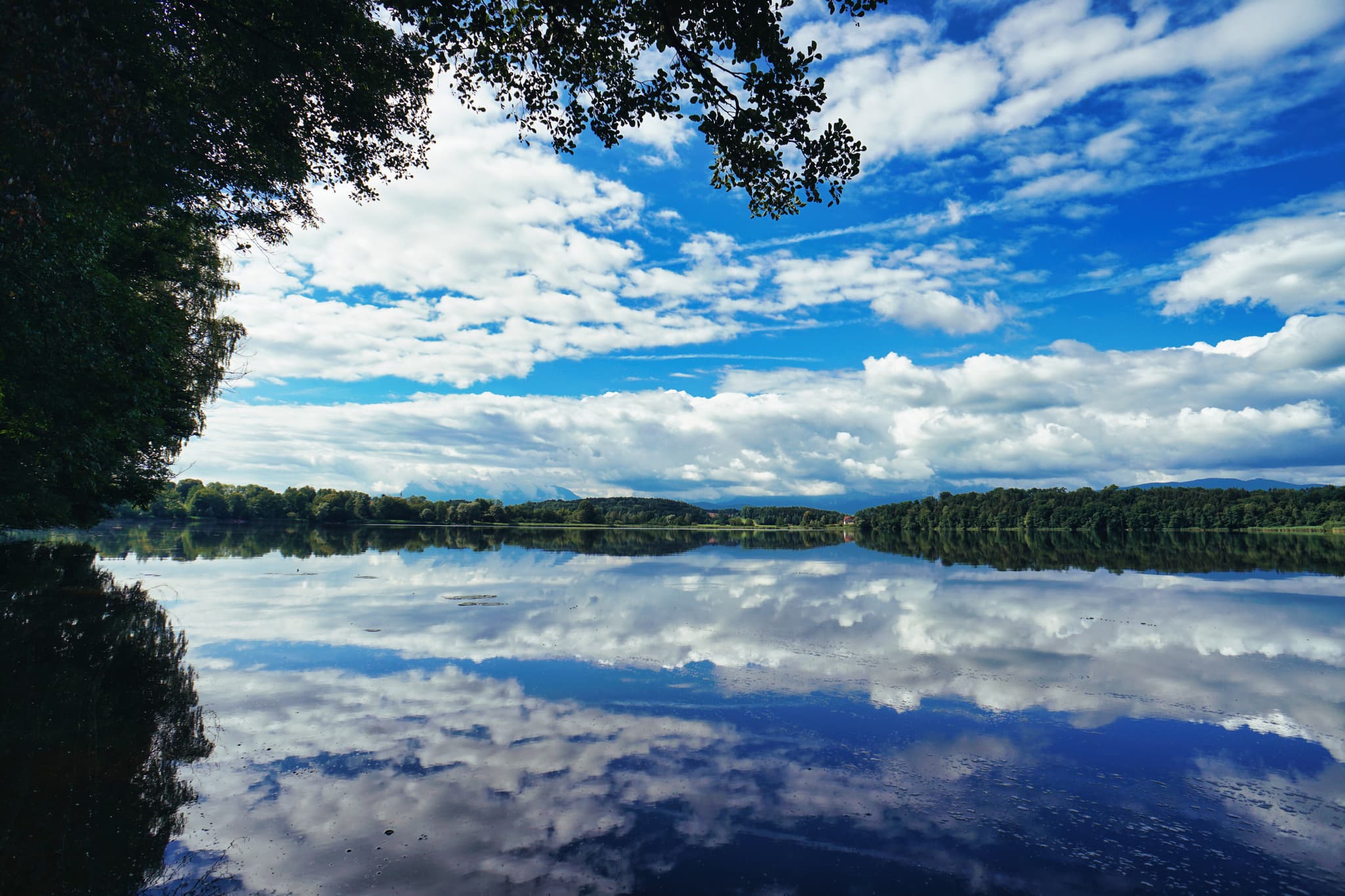 Ruhige Spiegelung des blauen Himmels mit weißen Wolken im Abtsee.  Die Ufer sind von Bäumen gesäumt, die im Vordergrund leicht im Schatten liegen.  