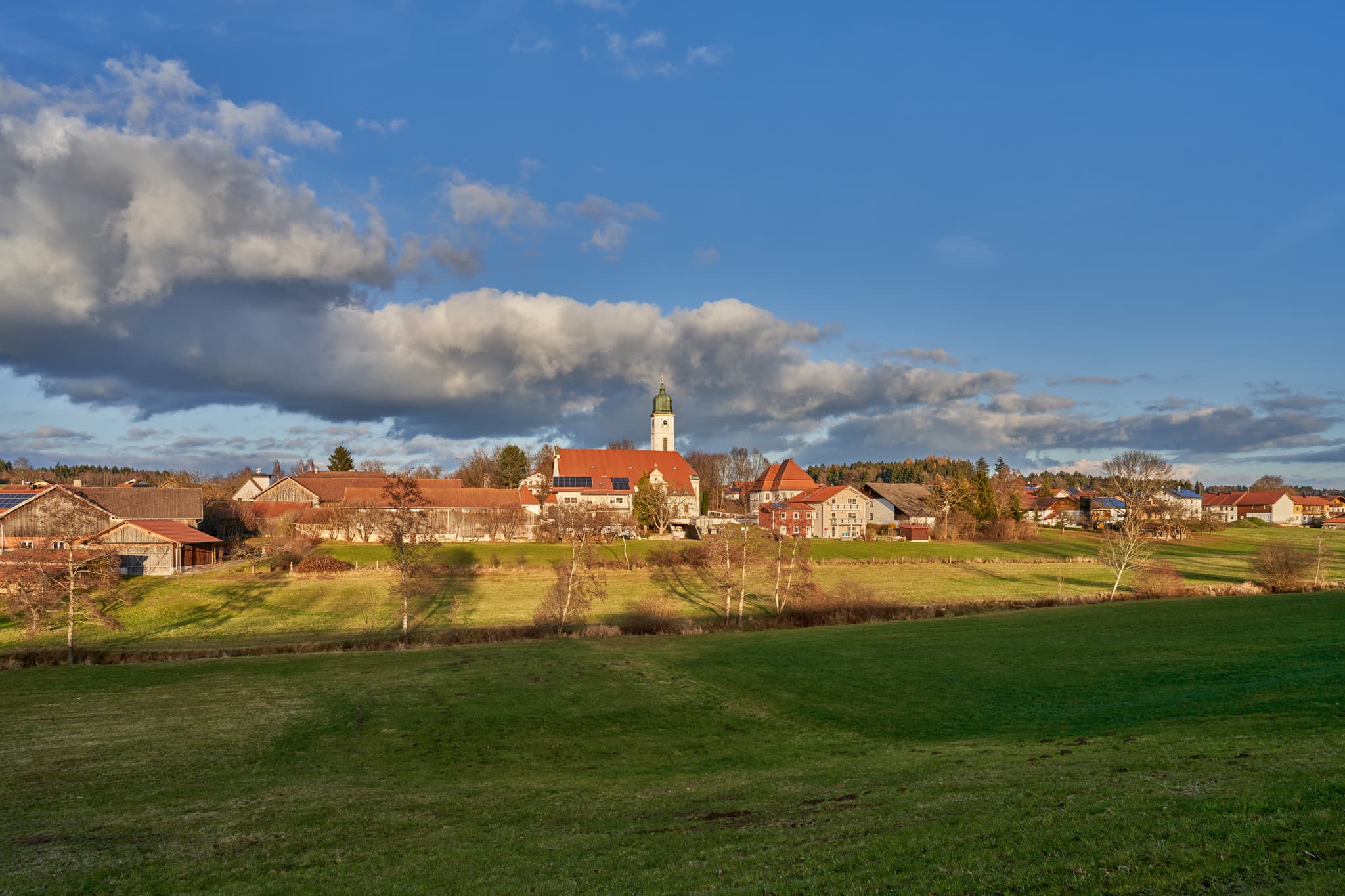 Ulbering, Wittibreut, Landkreis Rottal-Inn, Niederbayern. Ländliche Ansicht mit Kirche, Häusern, grünen Wiesen und Bäumen unter blauem Himmel.