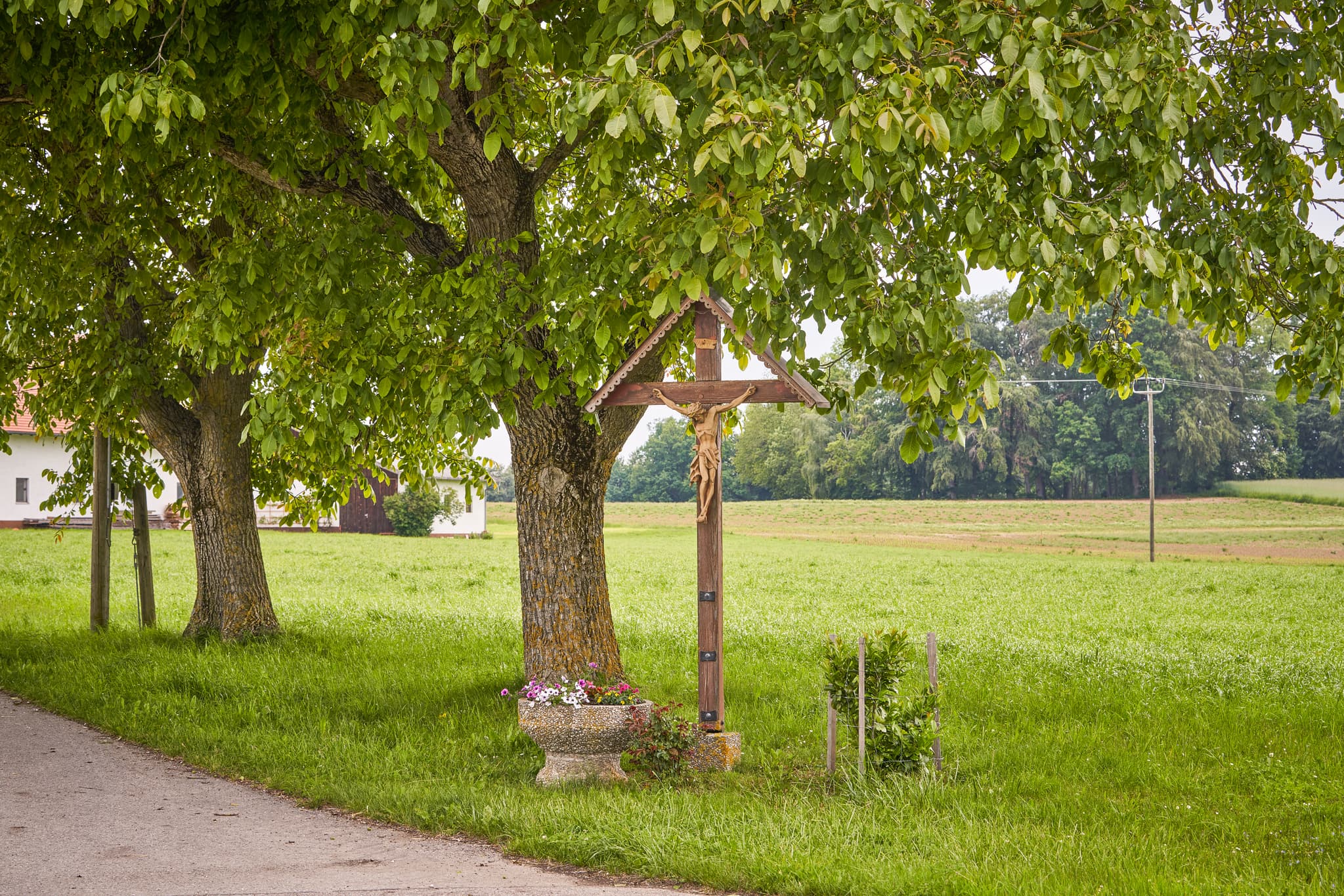 Wegkreuz unter Bäumen am Wegesrand in Bartlehen, Garching. Ländliche Szenerie in Altötting, Oberbayern, Deutschland. Teil der Region Inn-Salzach.