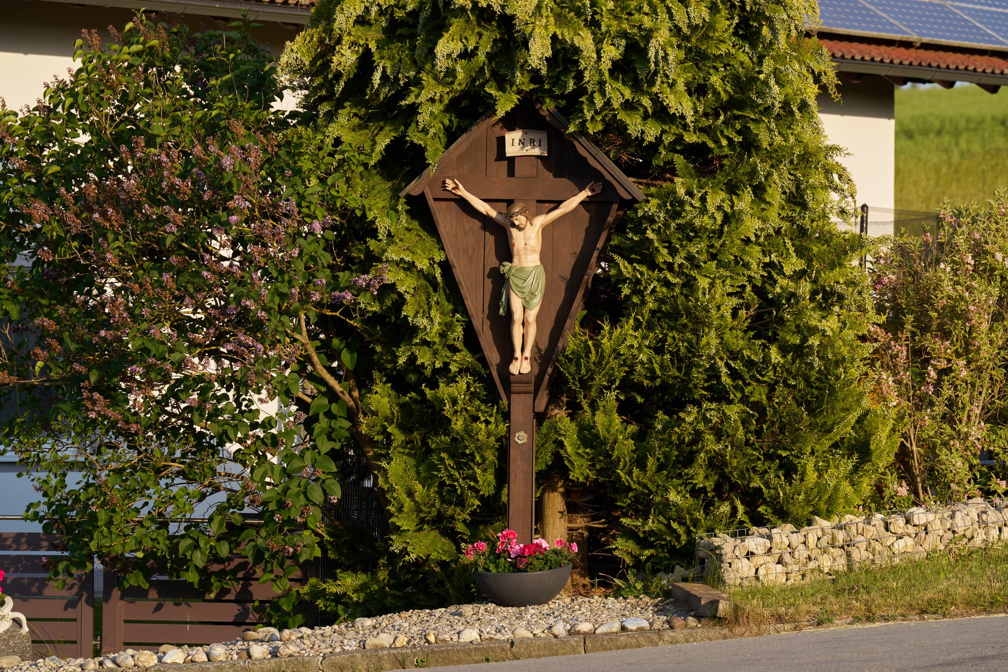 Hölzernes Wegkreuz an der Waldberger Straße in Arbing, Reischach, Altötting, Oberbayern. Umgeben von Sträuchern und Blumen.