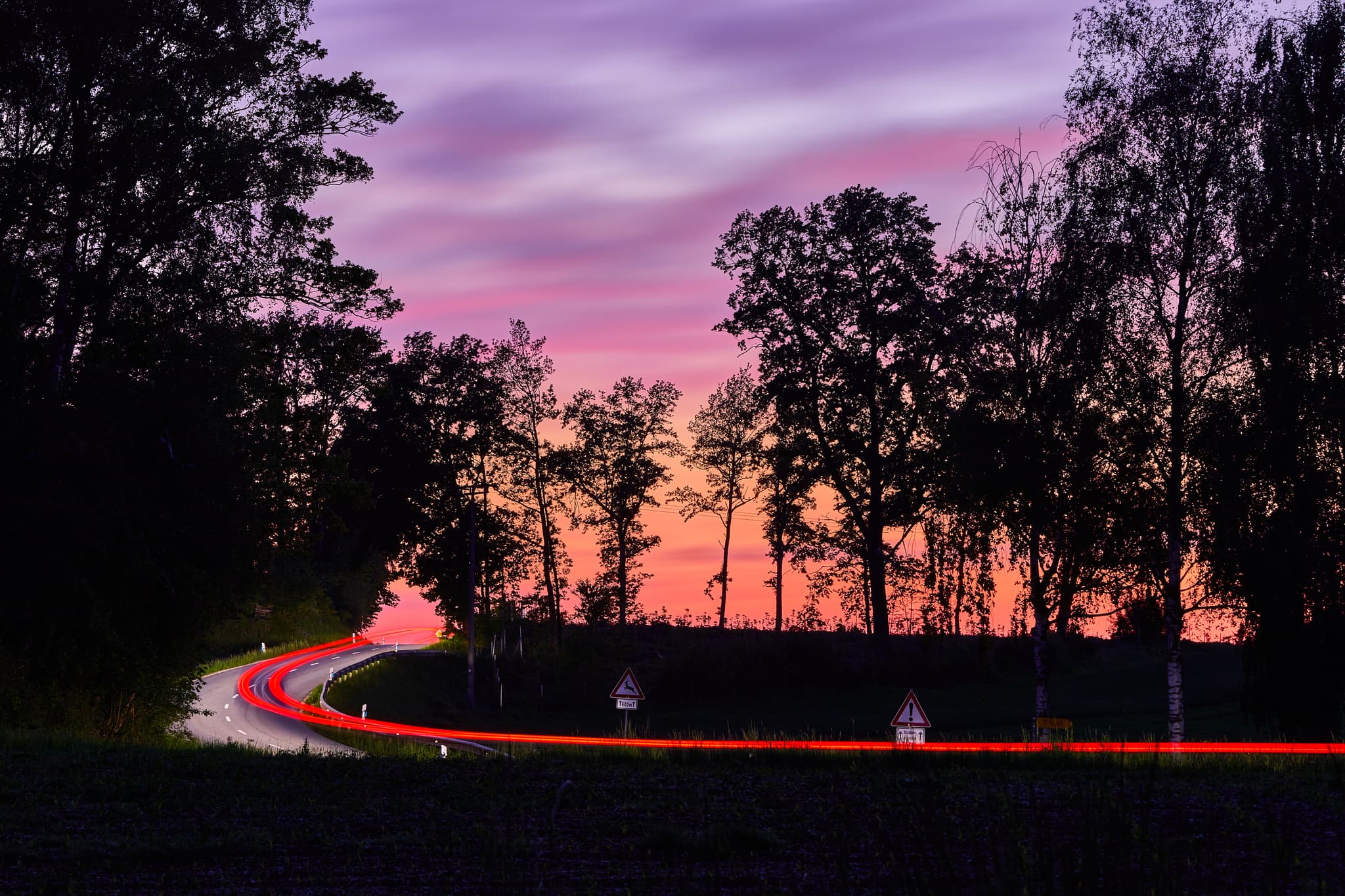 Nachtaufnahme einer kurvigen Straße bei Sonnenuntergang zwischen Wald und Georgenberg, nahe Pleiskirchen im Landkreis Altötting, Oberbayern
