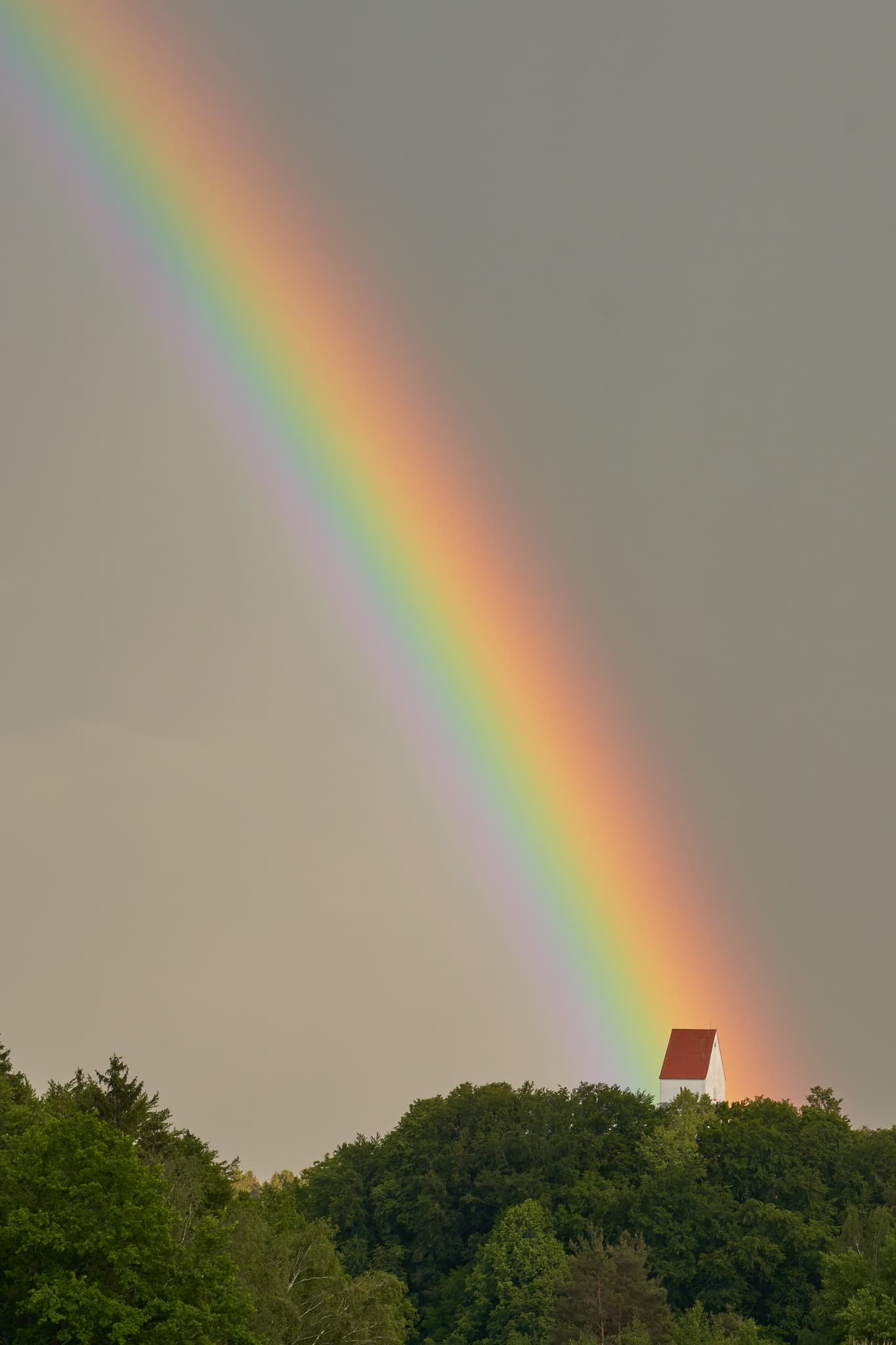 Farbenprächtiger Regenbogen über kleiner Kirche auf bewaldetem Hügel in Steinhausen, Erlbach, Altötting, Oberbayern, Inn-Salzach, Deutschland.
