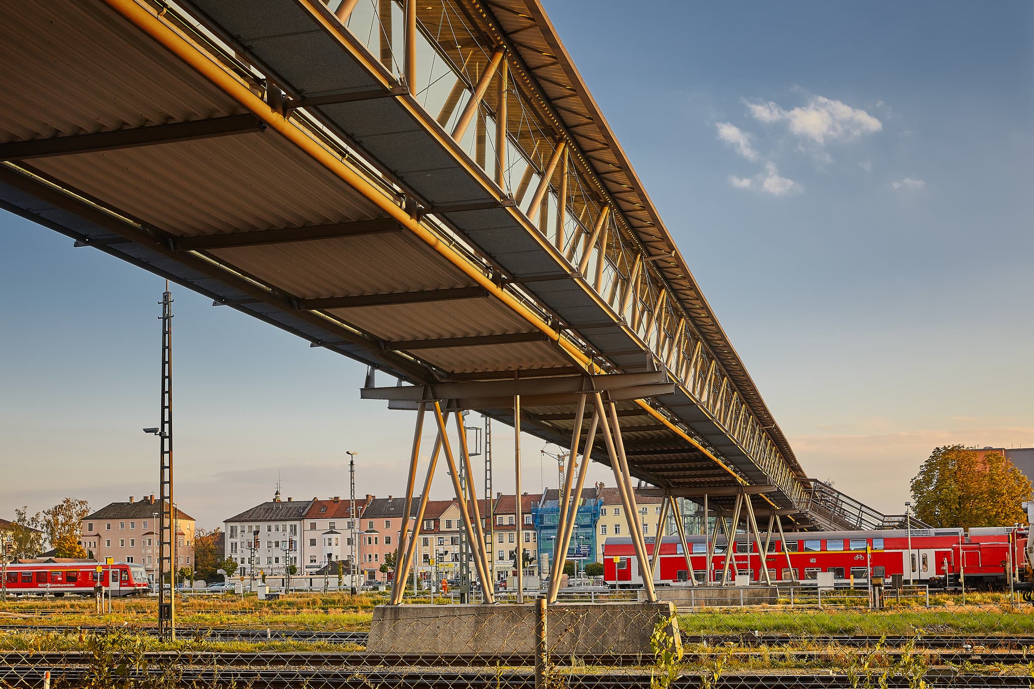 Fußgängerbrücke und Bahngleise am Bahnhof Mühldorf, Mühldorf am Inn, Oberbayern, Inn-Salzach, Deutschland. Rote Züge und Gebäude prägen die städtische Szenerie.
