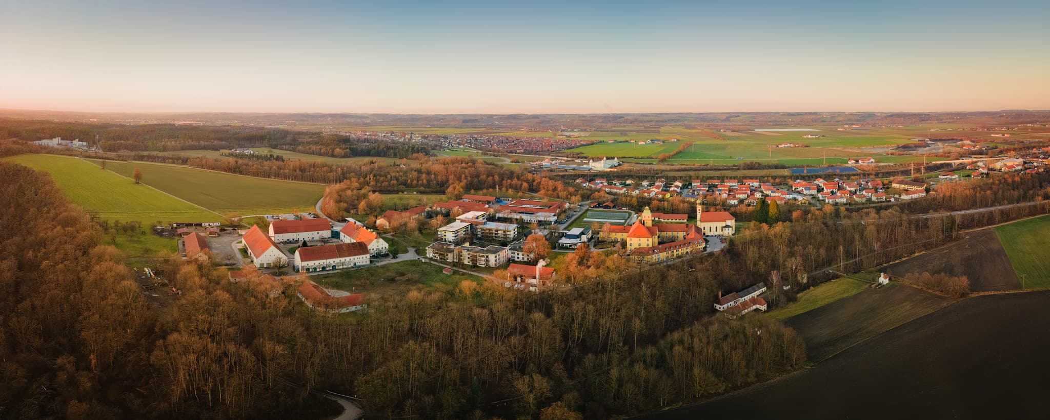 Luftaufnahme des Ortsteils Ecksberg, Mühldorf am Inn, Oberbayern. Zeigt Gebäudekomplexe, weite Wälder, Felder der Inn-Salzach Region in Abendstimmung.