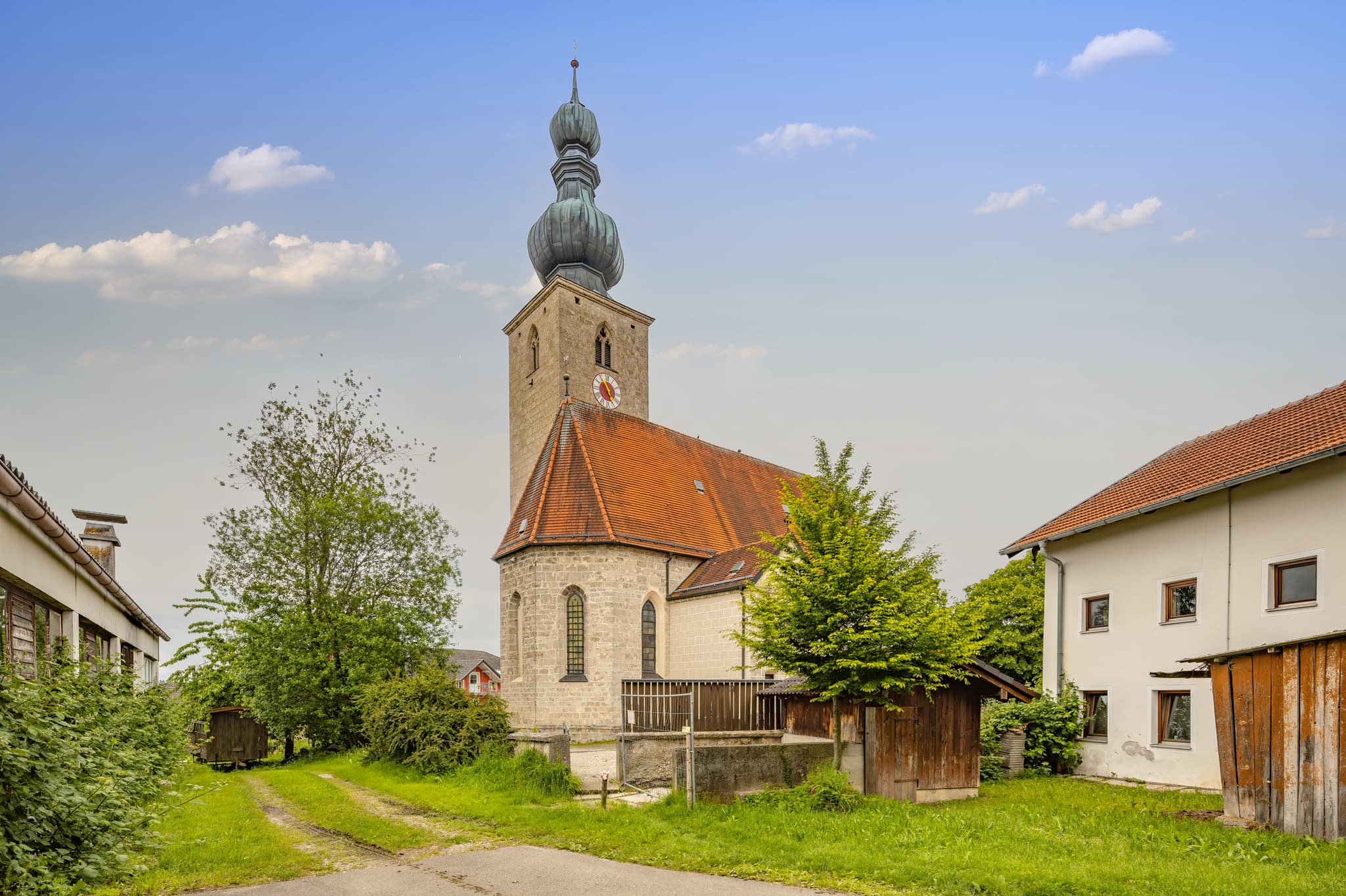 Die Pfarrkirche St. Johann Baptist in Tyrlaching, Landkreis Altötting, Oberbayern, Inn-Salzach, Deutschland. Ländlich gelegen, umgeben von Grün und Gebäuden.