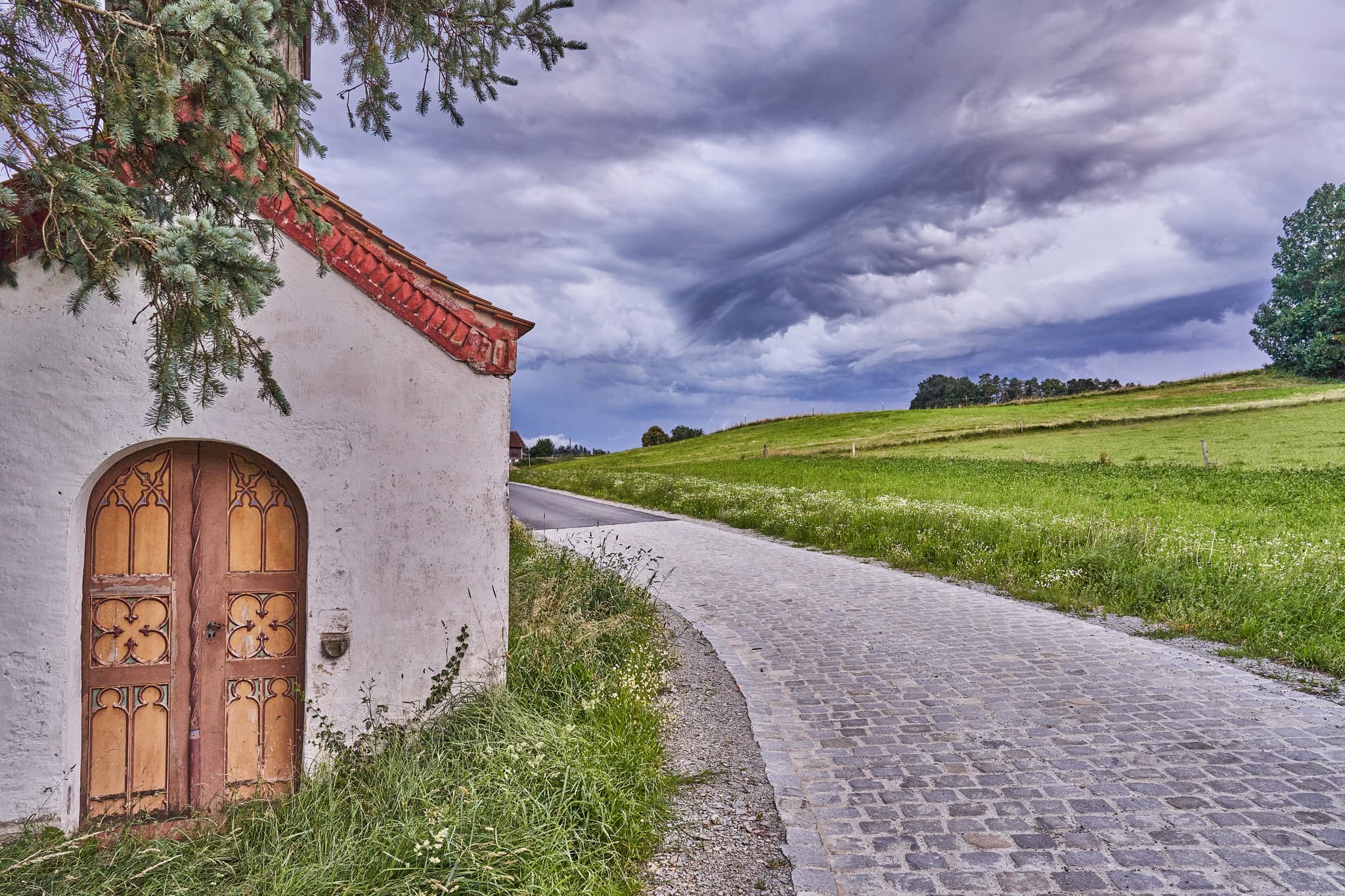 Perseis Kapelle, Oberthal, Reischach, Altötting. Kapelle, Kopfsteinpflasterweg, grüne Felder unter dramatischem Himmel. Inn-Salzach, Oberbayern, Deutschland.
