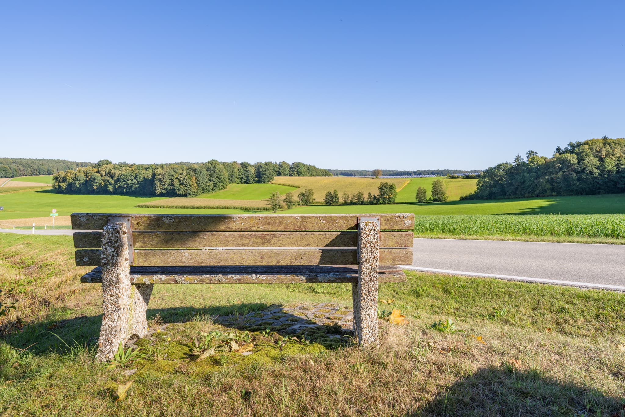 Sitzbank am Feldrand bei Baumgarten, Dietersburg im Landkreis Rottal-Inn, Niederbayern. Die Landschaft im Holzland, Deutschland, zeigt Maisfelder, Wälder.